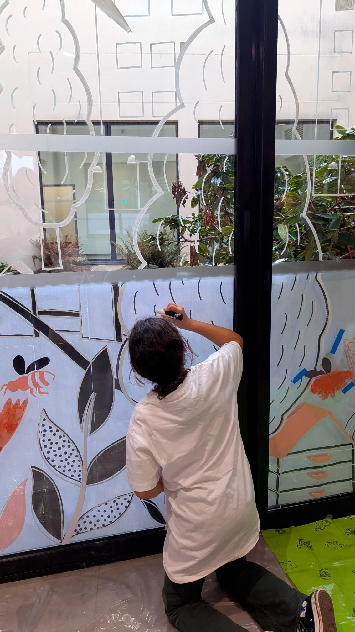 Fresque participative sur les vitrines du patio de la médiathèque des Mureaux, réalisée lors des Nuits de la lecture 2026. Thème "Villes et campagnes". Une jeune fille peint l'arbre de la fresque.