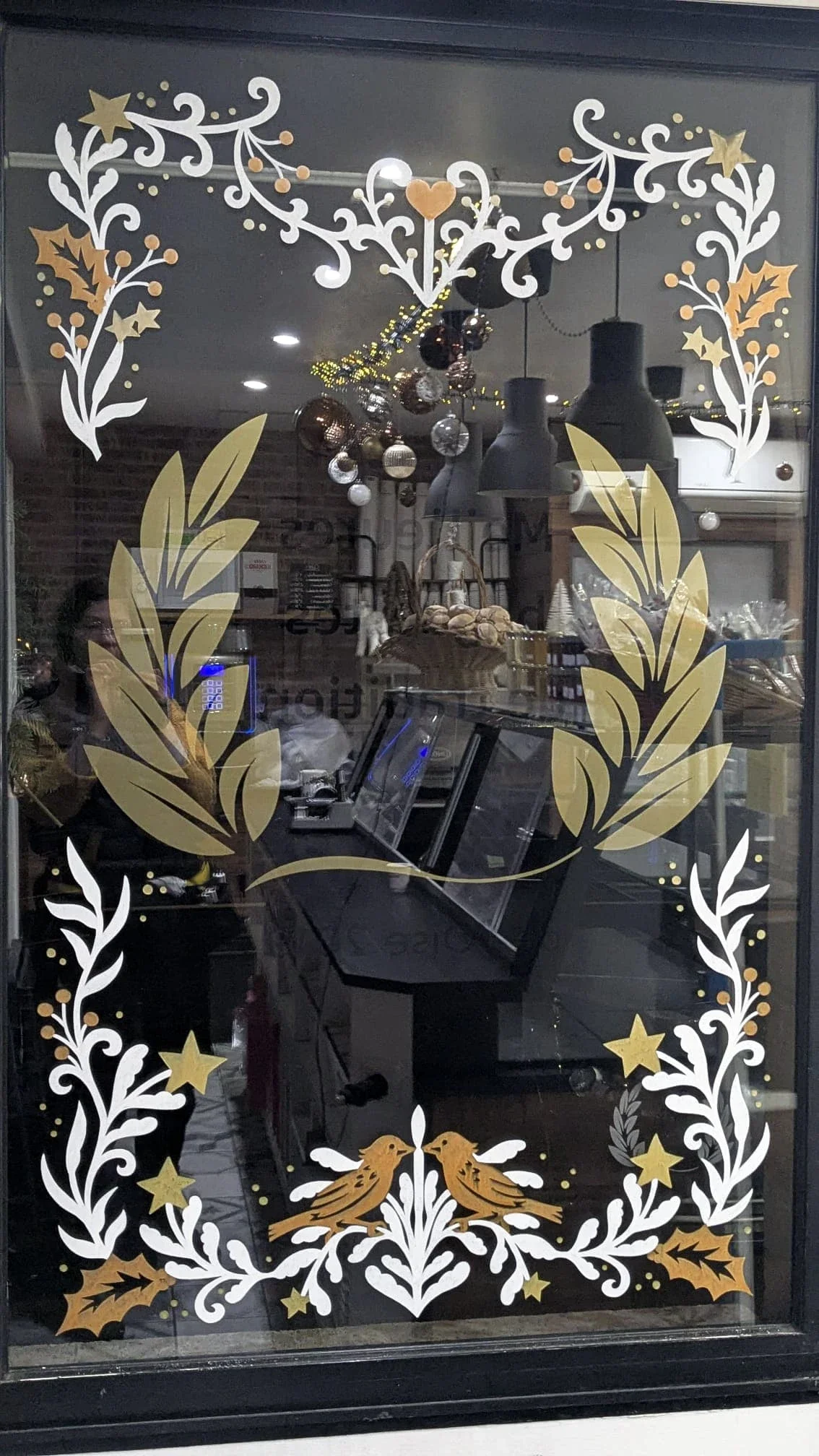 Peinture sur vitrine Noël pour une boulangerie à La Chapelle-en-Serval (60). Décor composé d'une frise avec végétaux, oiseaux, étoiles, baies et un coeur. Coloris blanc, cuivre et doré