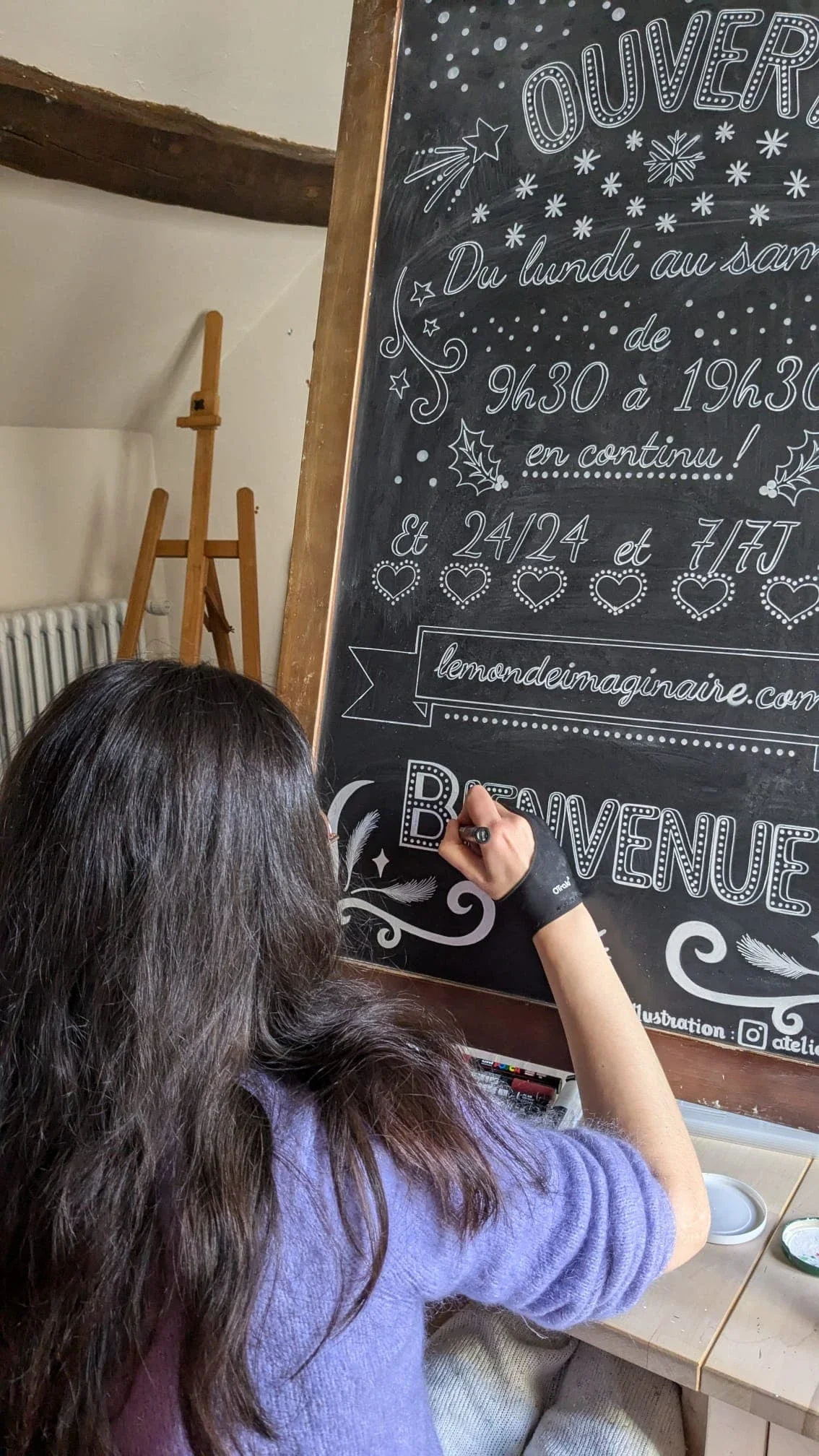 Sandrine Daniel peint à la main le mot « Bienvenue » sur l'ardoise stop-trottoir d'une boutique de jouets dans son atelier de Vaux-sur-Seine