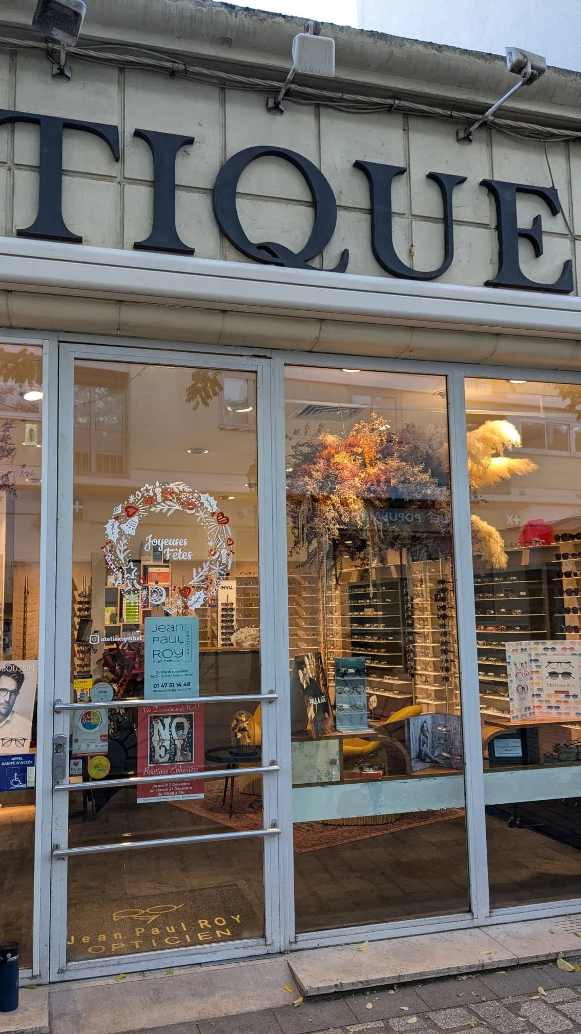 Peinture sur vitrine représentant une couronne de Noël peinte à la main avec houx, cloches, étoiles, baies et branches de pin pour l'opticien Jean-Paul-Roy à Rueil-Malmaison (92) Plan large sur la devanture de la boutique.