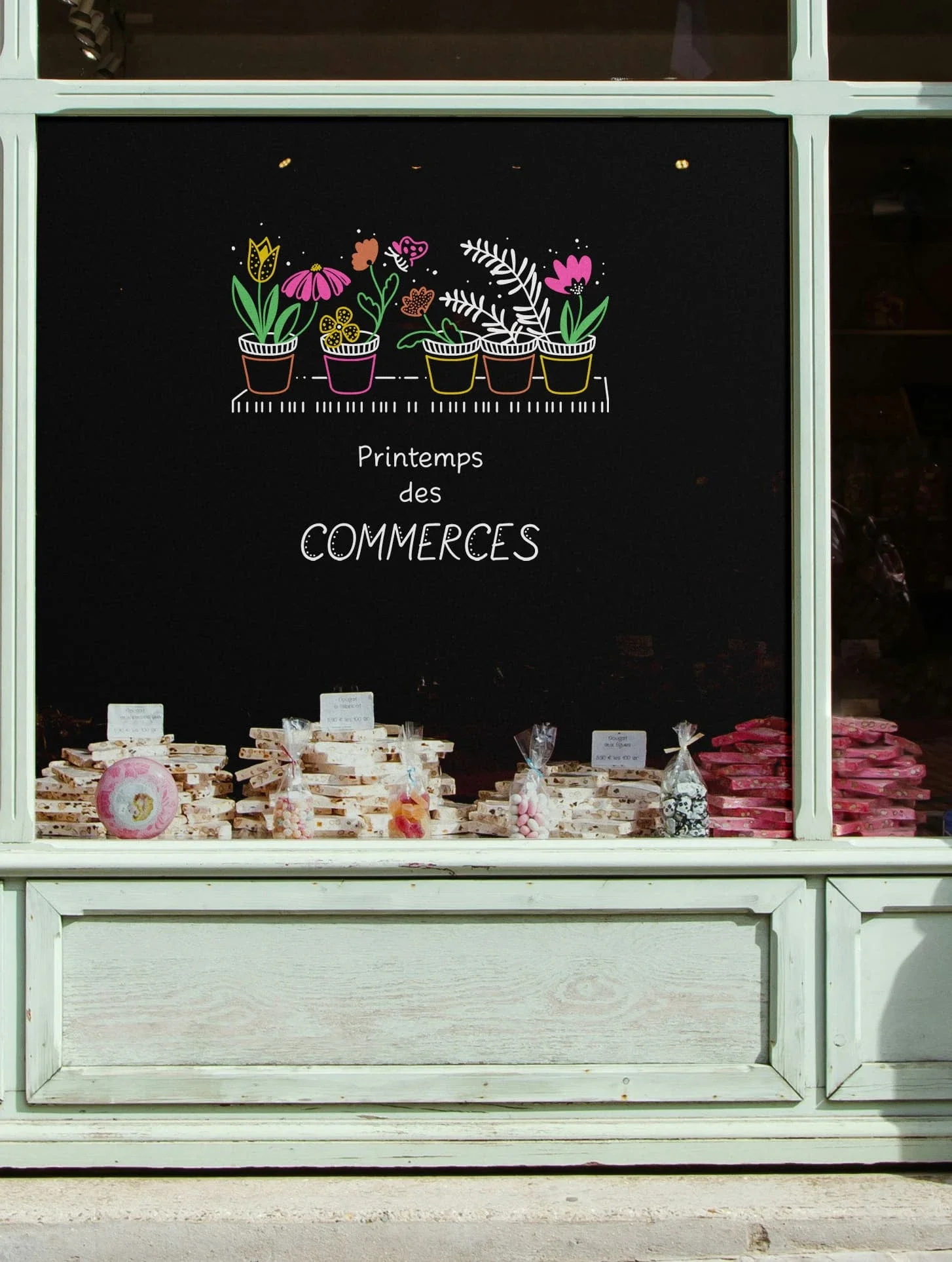 Peinture sur vitrine sur la devanture d'une boutique. Thématique "Printemps des commerces". Pots de fleurs sur une étagère. Lettrage blanc. Coloris blanc, vert, rose, jaune et orange