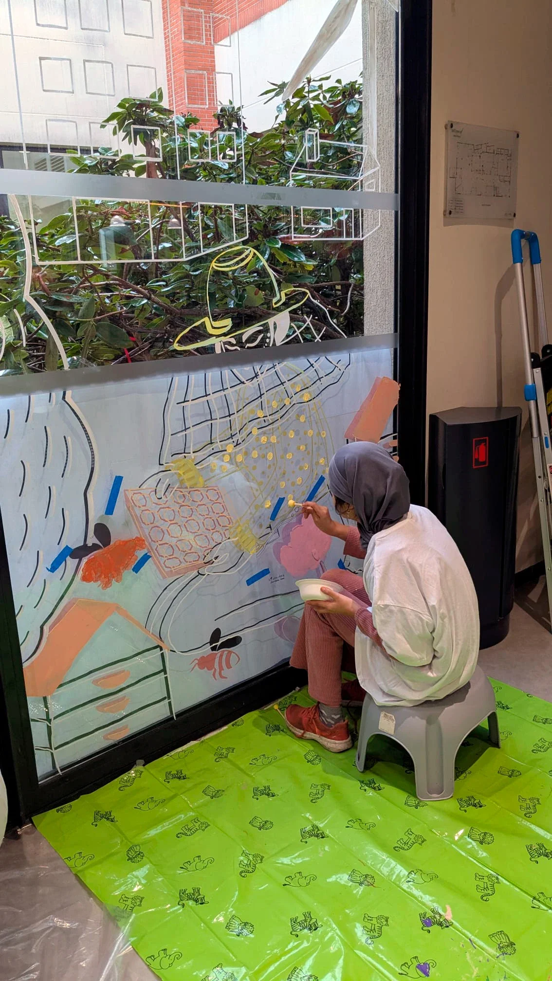 Fresque participative sur les vitrines du patio de la médiathèque des Mureaux, réalisée lors des Nuits de la lecture 2026. Thème "Villes et campagnes". Une jeune femme peint avec un tampon les motifs pois de l'habit du personnage. Peinture acrylique 