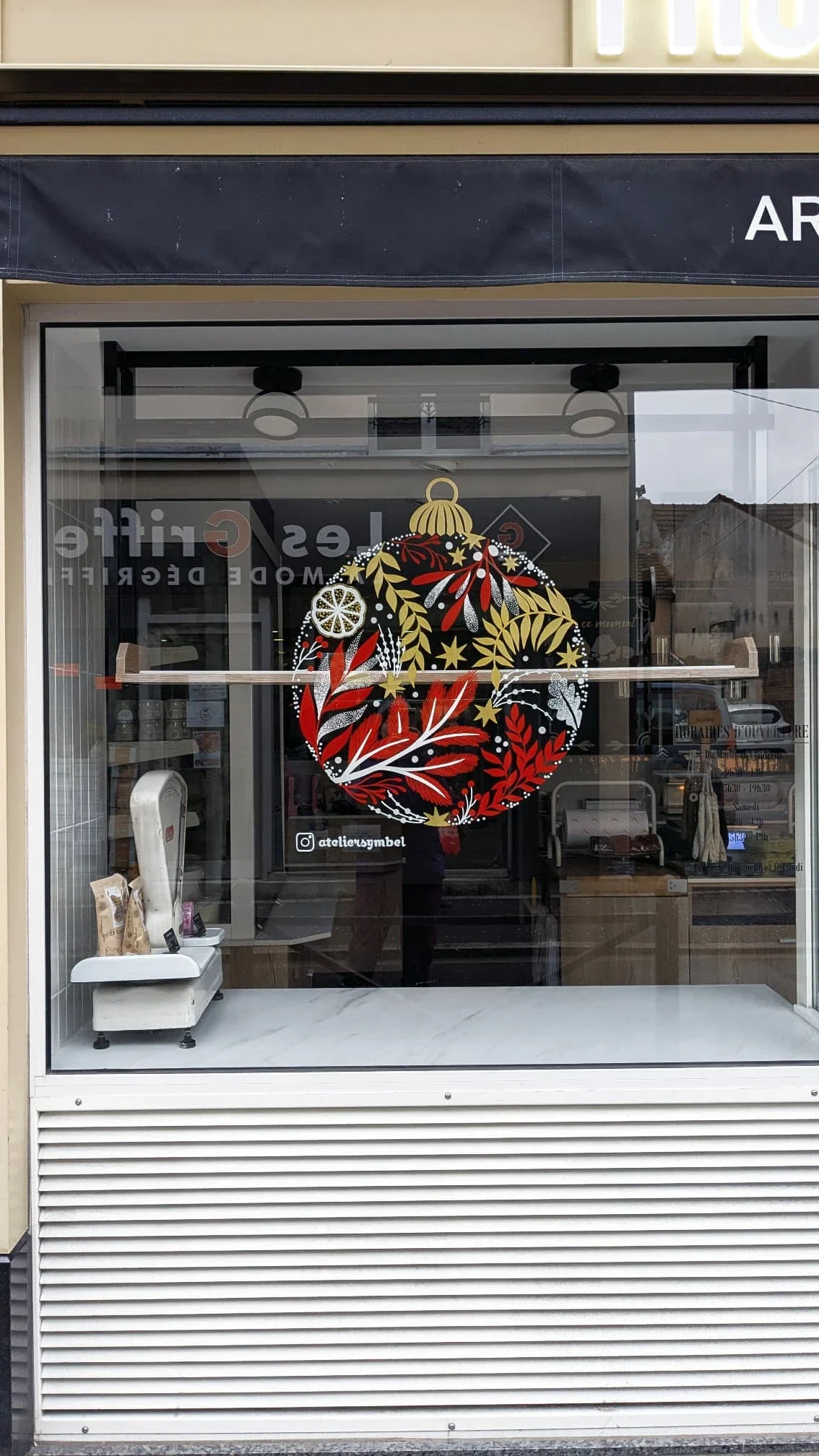 Vue rapprochée de la peinture sur vitrine de Noël réalisée à la main pour la fromagerie Lallias à Houilles, représentant une boule de Noël décorée de végétaux. coloris blanc, rouge et doré