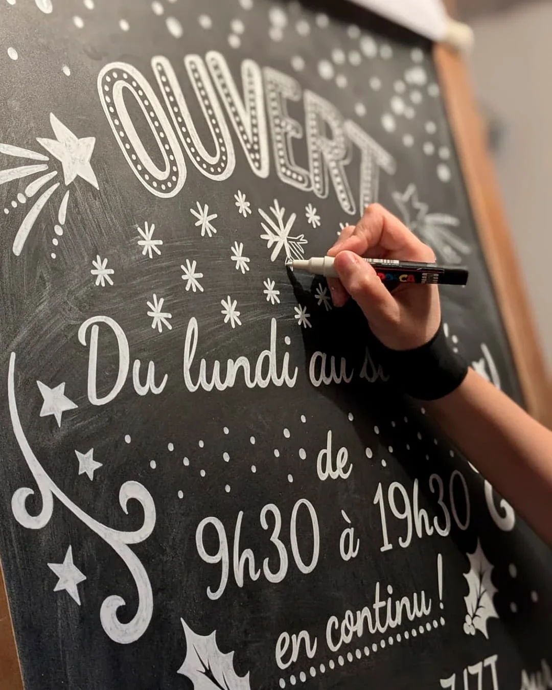 Ardoise illustrée, peinte à la main à l'acrylique blanche. Lettrages informant sur les horaires d'ouverture. Décor d'étoiles, d'entrelacs et de points