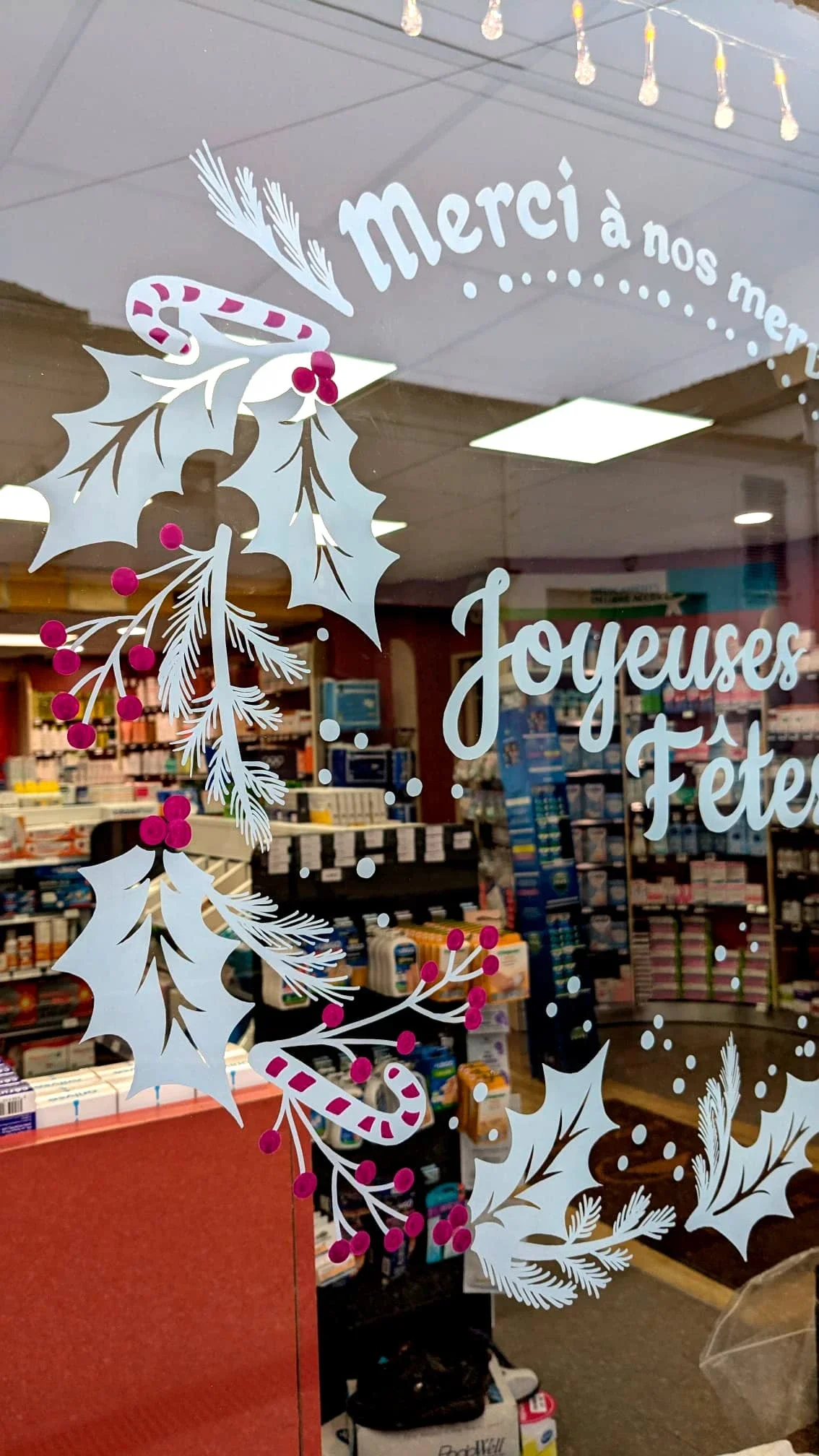 Gros plan sur la peinture sur vitrine de Noël représentant une couronne Houx et sucre de canne réalisée pour la pharmacie du Précoquet à Vaux-sur-Seine (78). Lettrage Joyeuses fêtes. COloris rose et blanc