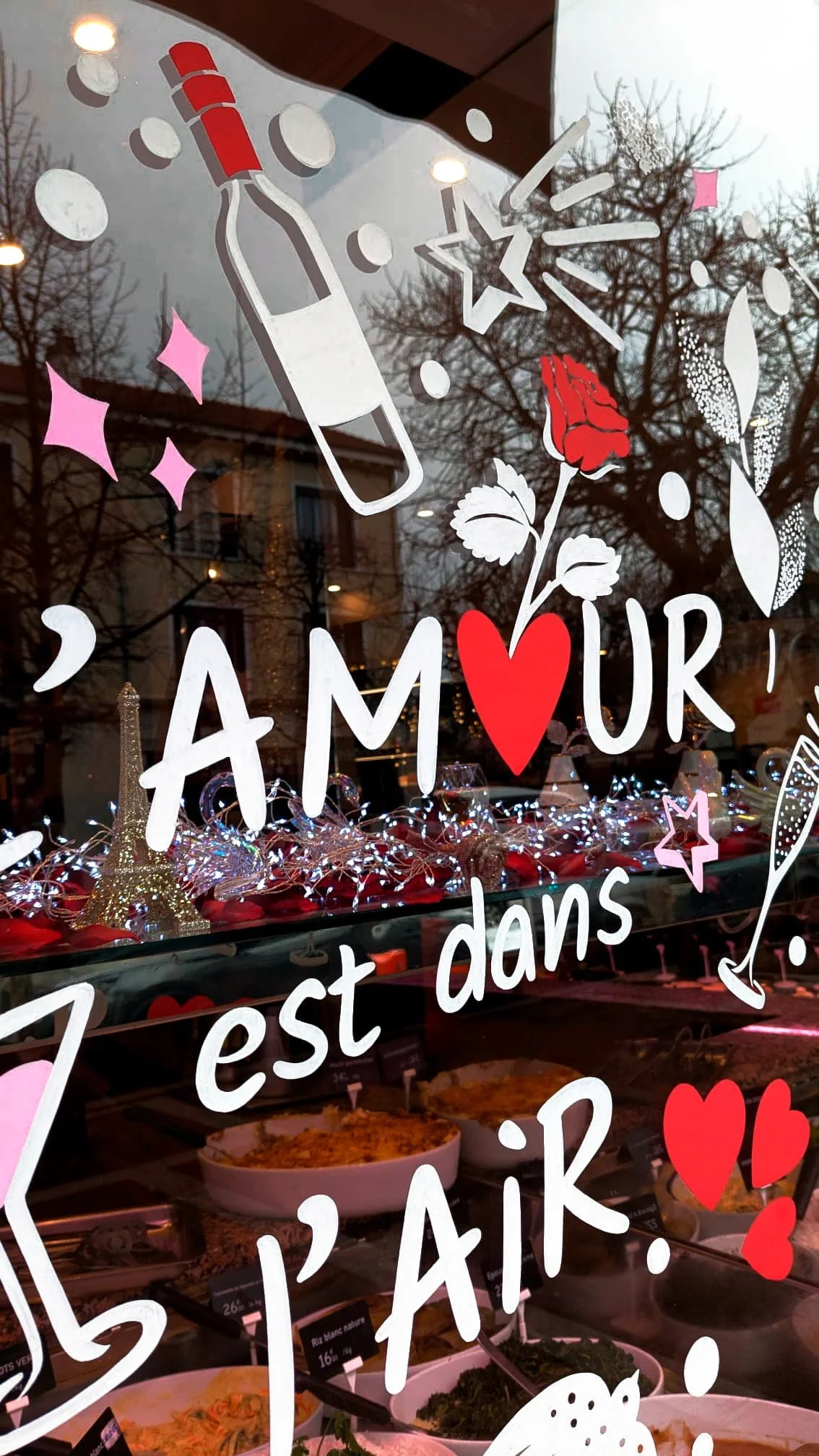 Peinture artisanale sur vitrine pour la Saint-Valentin avec lettrage peint, motifs de roses, cœurs, étoiles, bouteille de vin et verre à champagne, coloris blanc, rouge et rose, chez un traiteur à Croissy-sur-Seine.