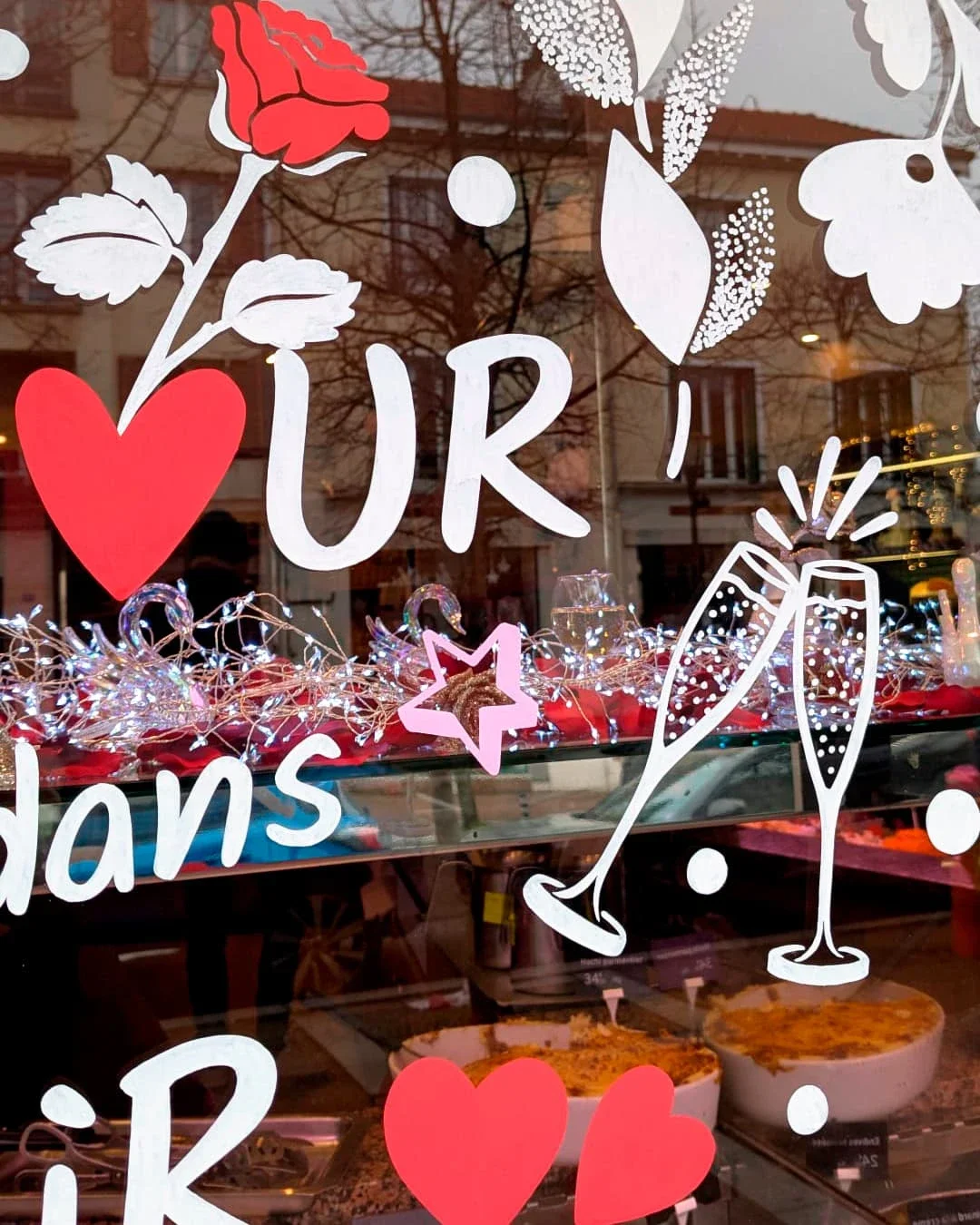Peinture sur vitrine,  thématique Saint-Valentin, gros plan sur les verres de champagne, la rose, les cœurs et les éléments floraux. Coloris blanc, rouge et rose