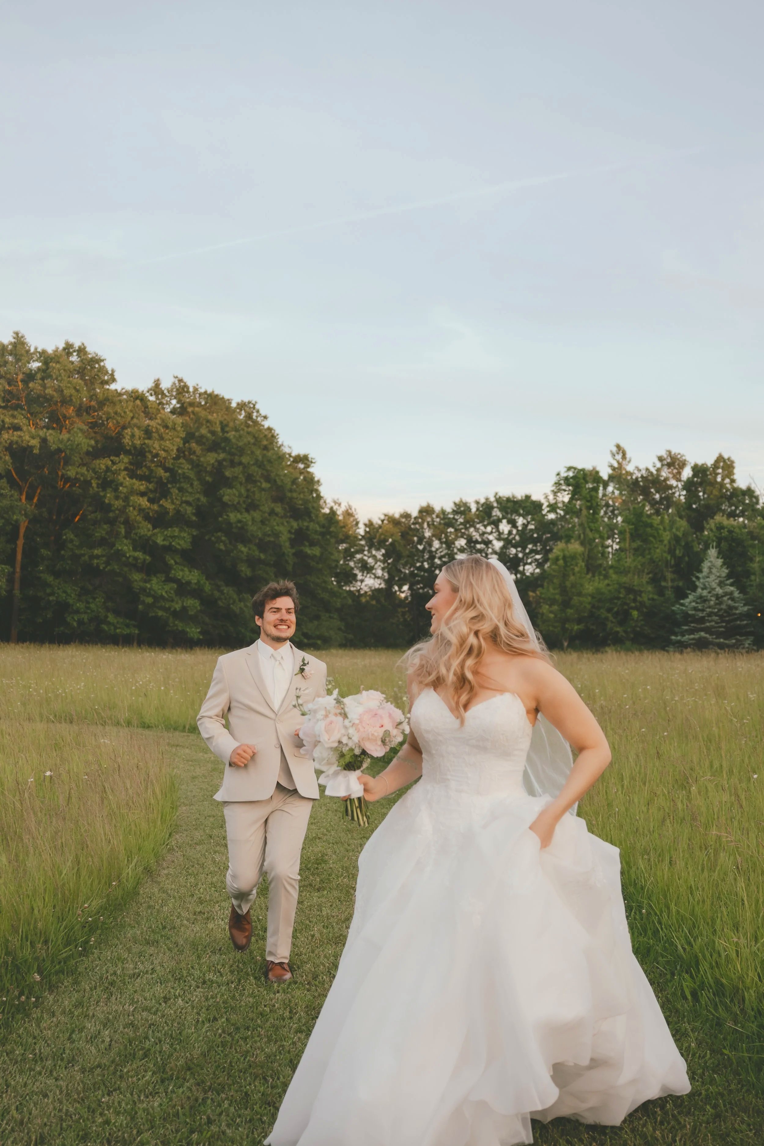 A wedding scene in an open field with a bride in a white wedding gown holding a bouquet, and a groom in a beige suit walking behind her. There are trees in the background and a clear sky overhead.