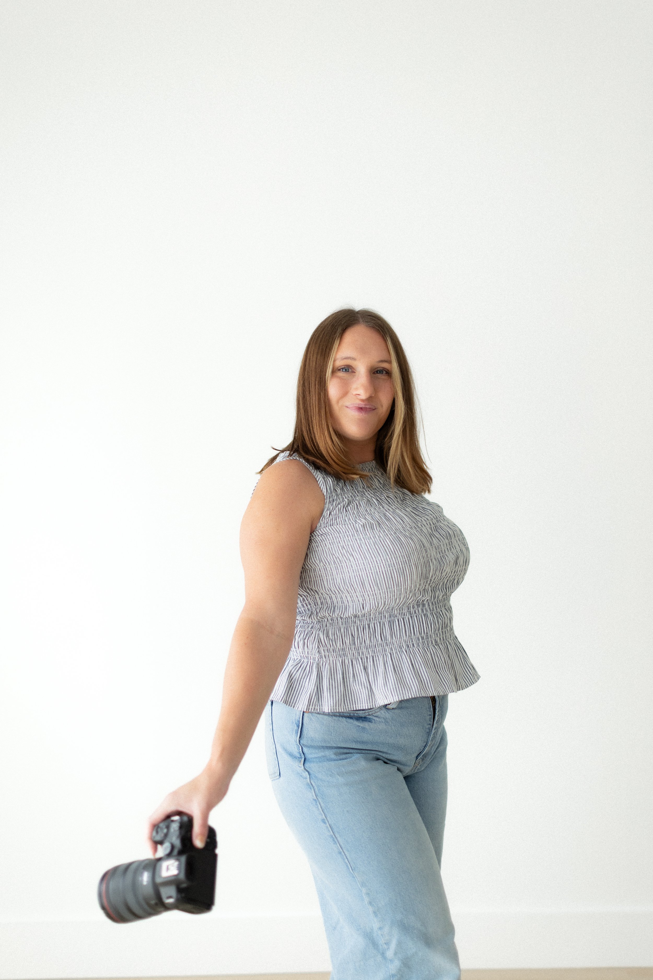 A woman with brown hair, wearing a sleeveless, striped top and jeans, is holding a camera with a zoom lens in her right hand against a plain white background.