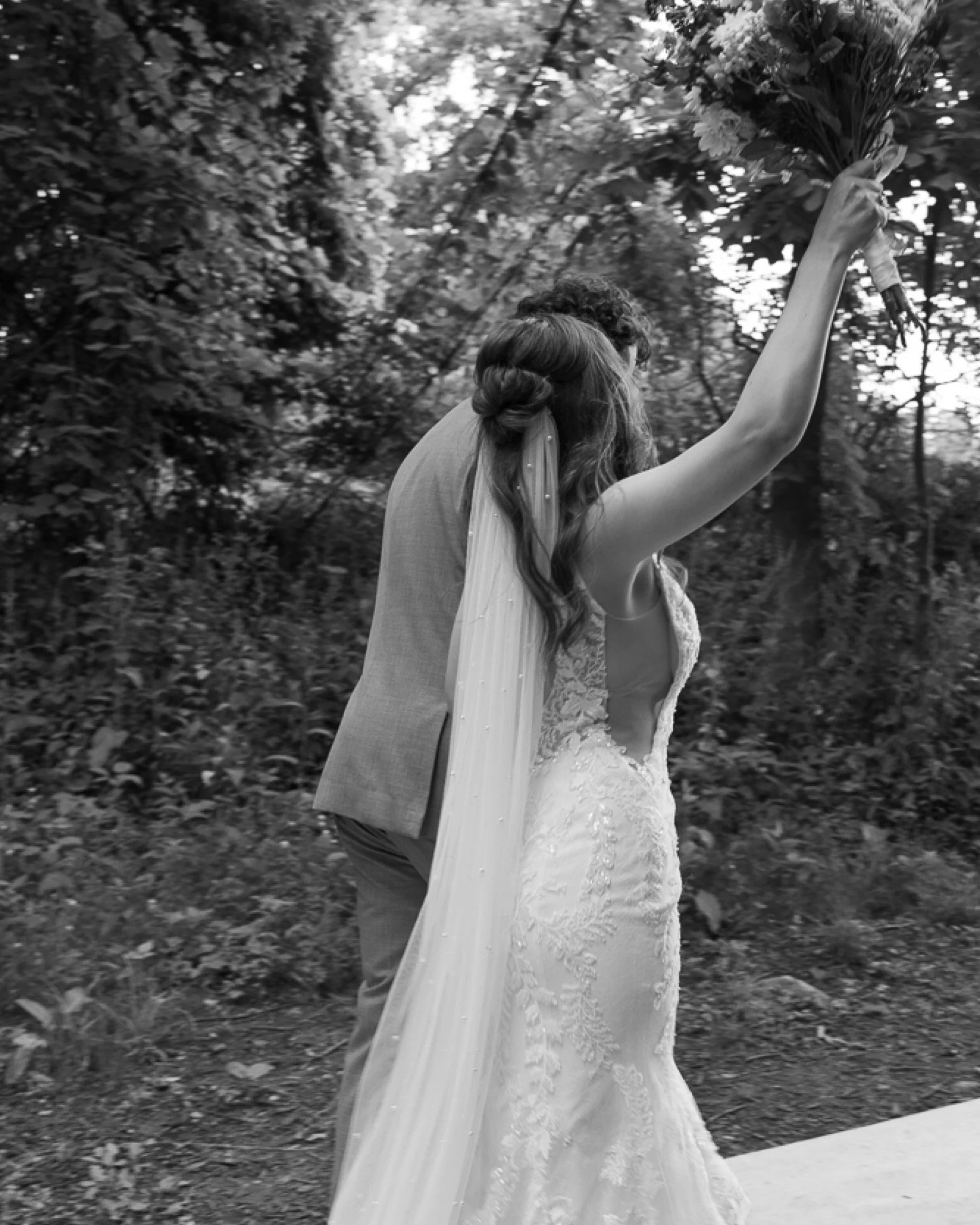 A bride and groom walking outdoors, the bride holding a bouquet of flowers, with trees in the background.