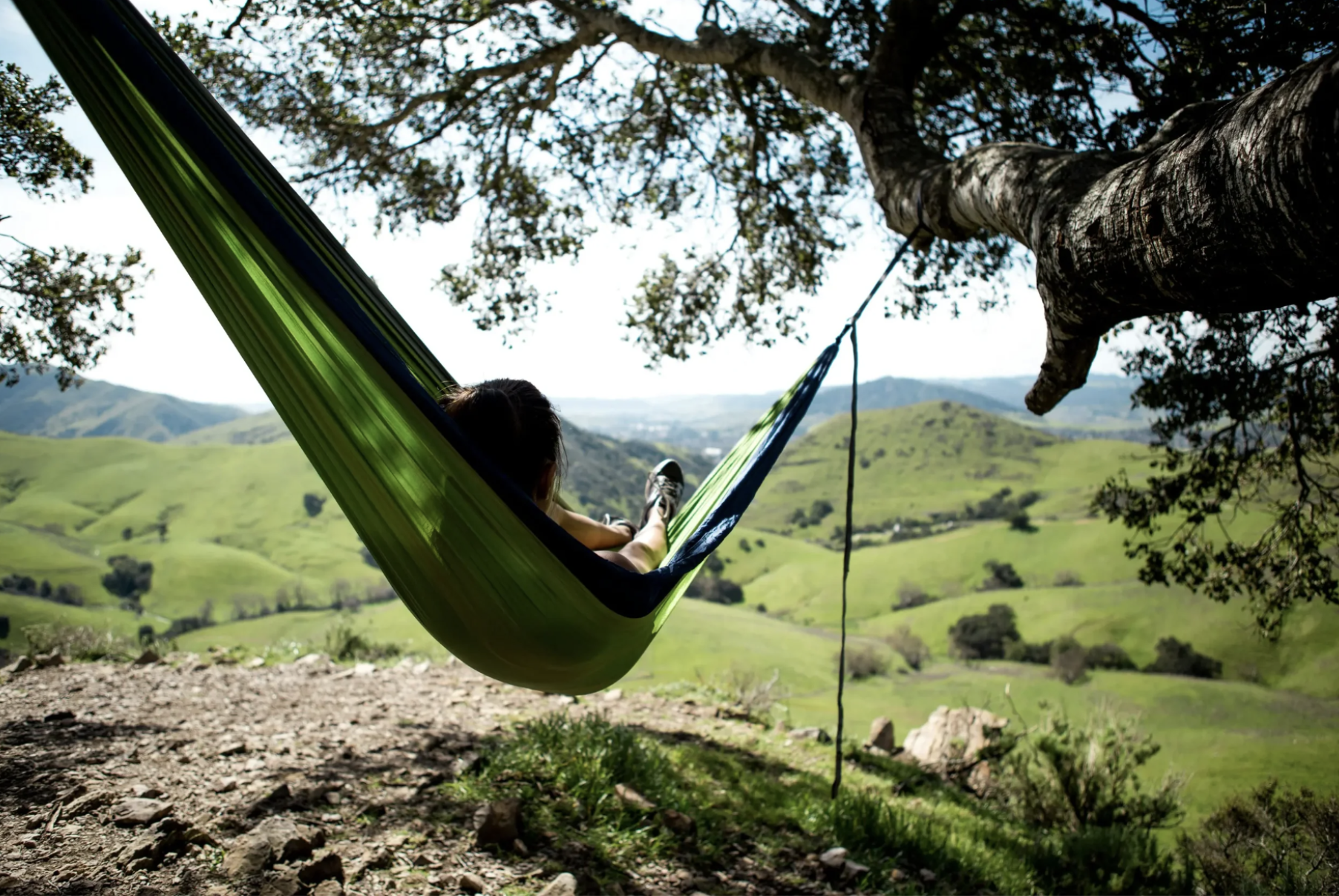 Person relaxing in a lime green hammock tied to a large tree, overlooking green rolling hills and distant mountains on a sunny day.