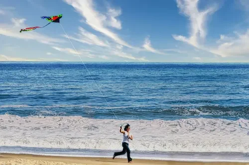 A woman flying a colorful kite on the beach with ocean waves in the background.