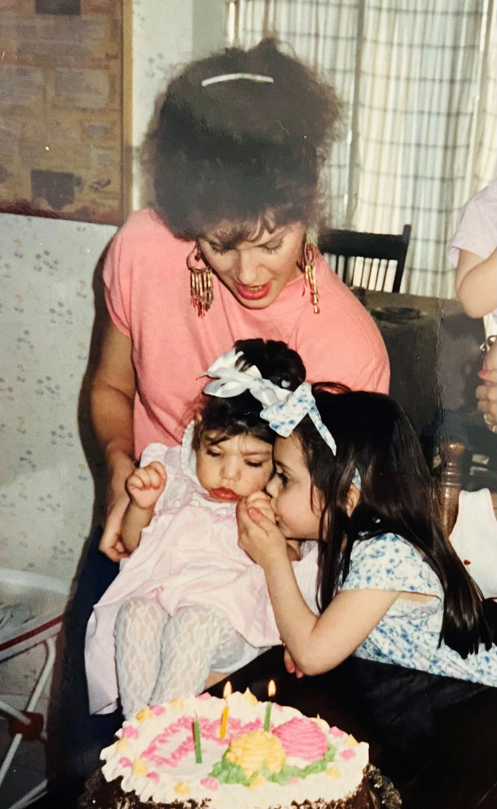 A woman with dark hair and earrings, wearing a pink top, is leaning over two young girls sitting in front of a birthday cake with lit candles. One girl is holding a hand near her mouth, while the other is whispering into her ear. The girl sitting in 
