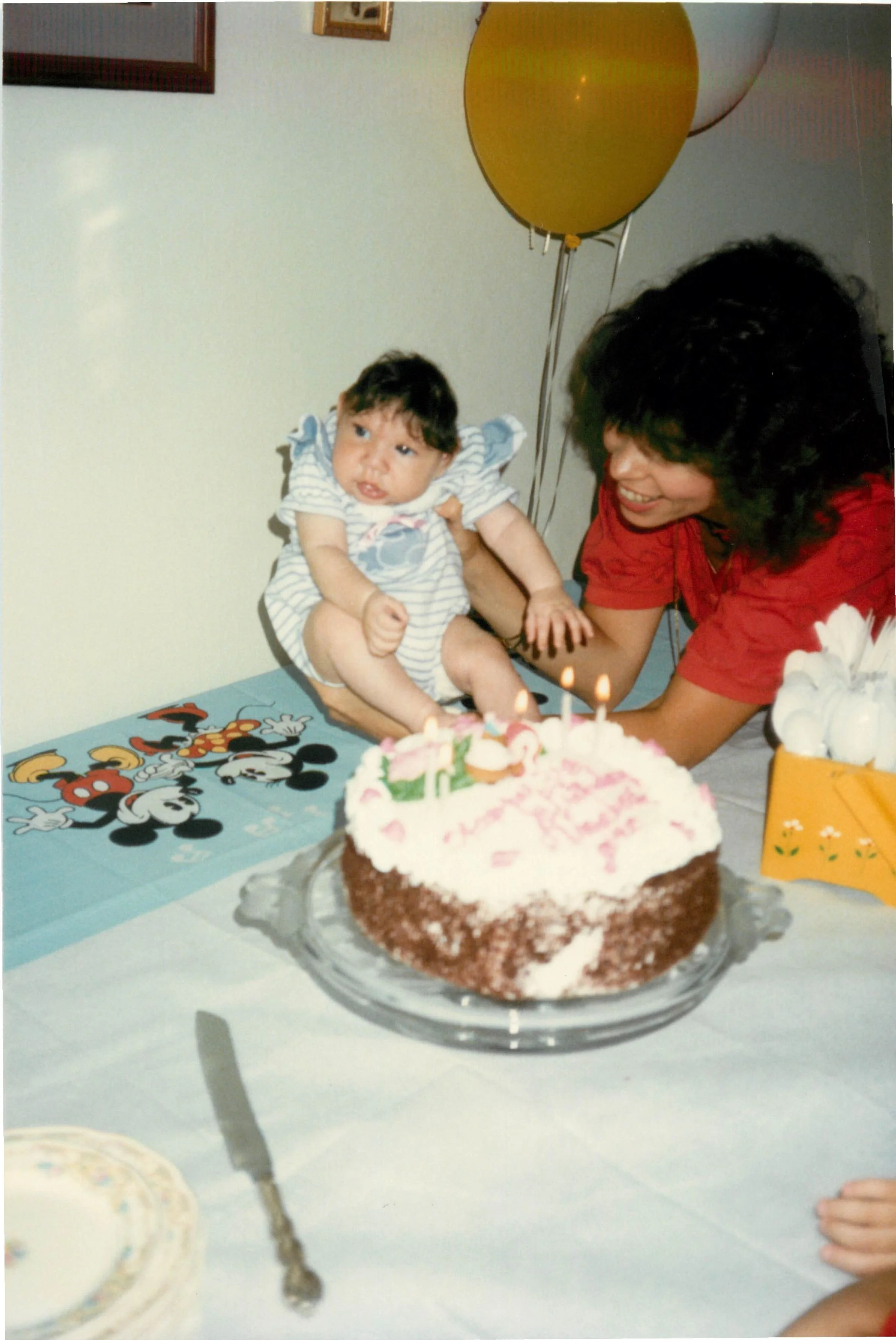 A woman in a red shirt holding a young child at a birthday celebration. The child is sitting on the table near a birthday cake with pink and white frosting and lit candles. There are two yellow balloons in the background, and the table has a Mickey M