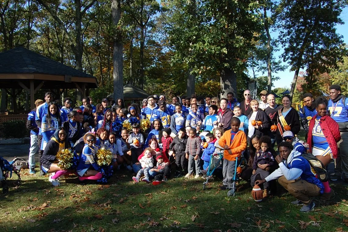 A large group of people including children and adults, in an outdoor park, some dressed in cheerleading uniforms, some with sports equipment, posing for a group photo among trees with autumn leaves.