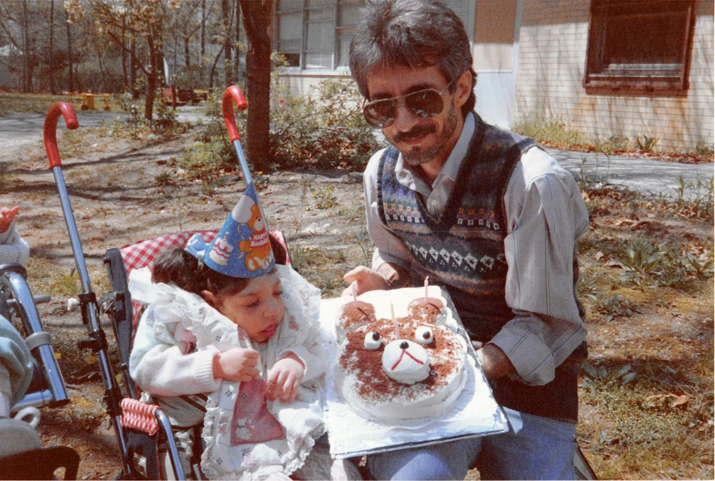 A young girl in a wheelchair wearing a birthday hat, sitting outdoors with a man holding a birthday cake with a bear face decoration. The man is smiling and wearing sunglasses, and the birthday hat has a bear illustration. The background shows trees 
