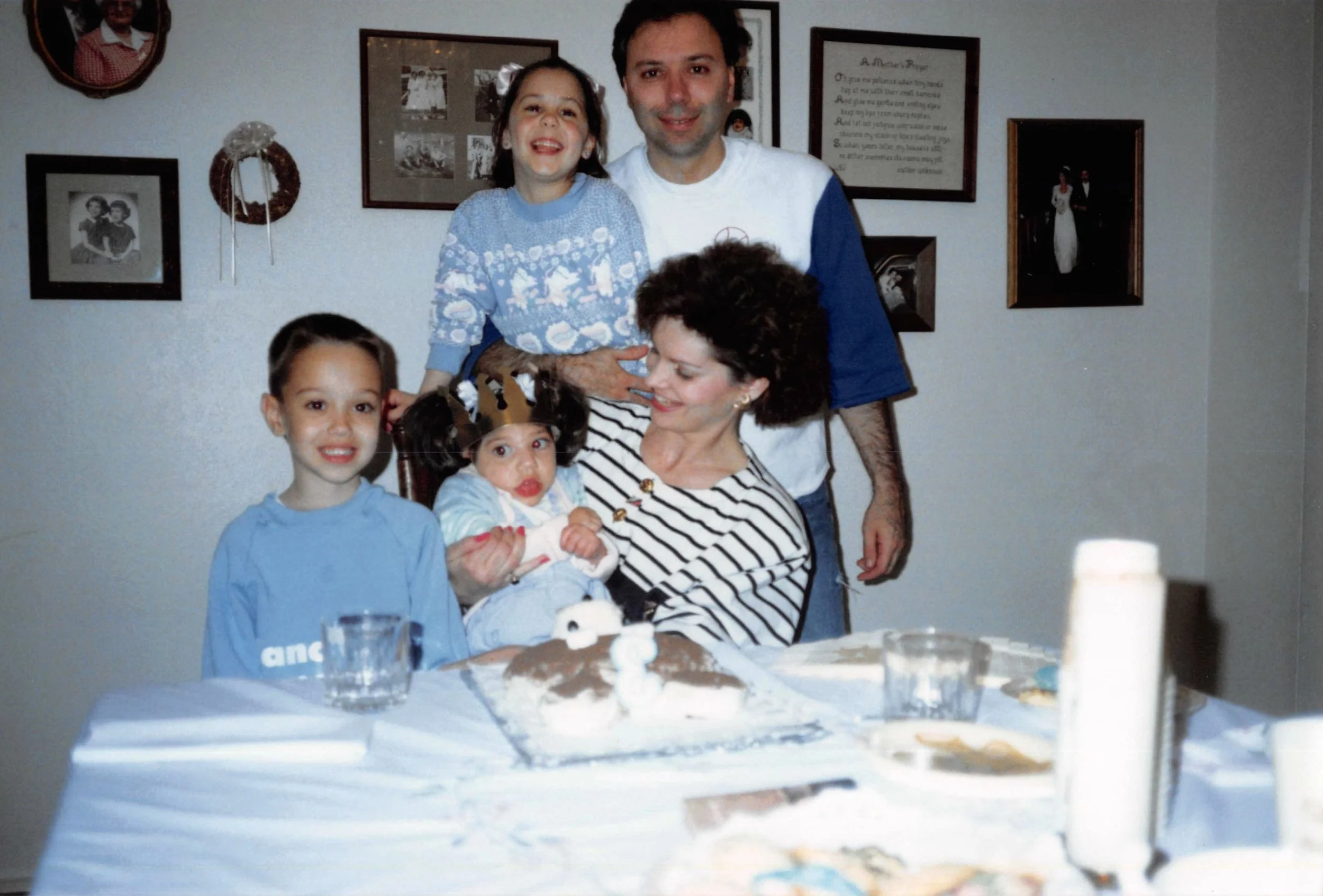 A family gathers for a birthday celebration in a home, with a birthday cake on the table and five family members smiling. There are framed photos and decorations on the wall behind them.