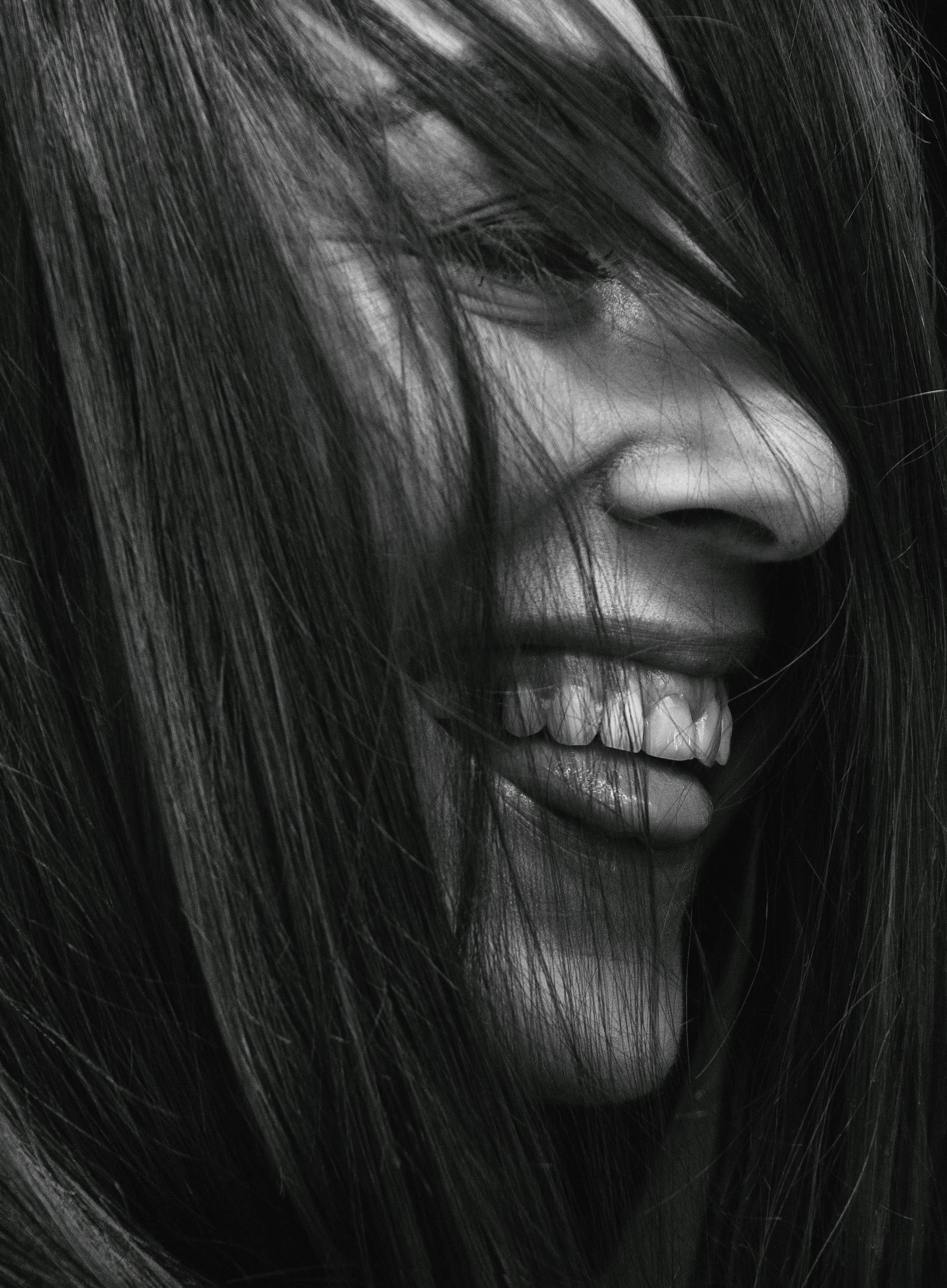 Close-up black-and-white photo of a woman with long, dark hair covering part of her face, smiling slightly with visible teeth.