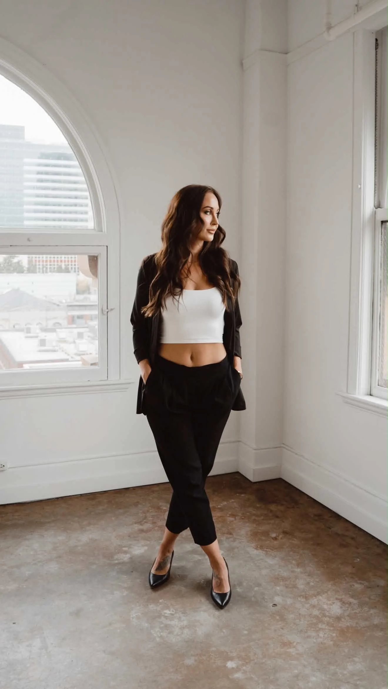 A woman with long dark hair standing in a room with white walls and large windows, wearing black pants, a white crop top, a black blazer, and black high heels.