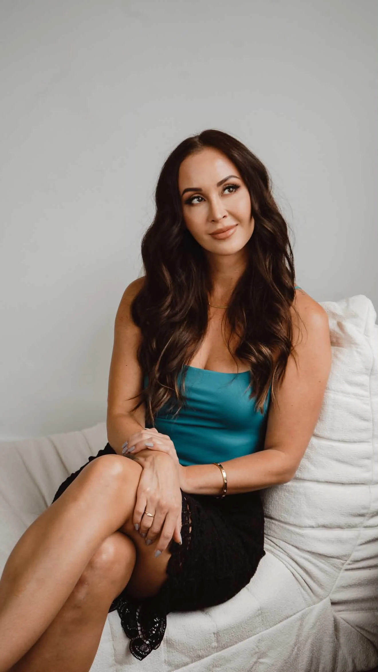 A woman with long, dark wavy hair wearing a teal top and black lace shorts sitting on a white couch, looking to her right with a neutral expression.