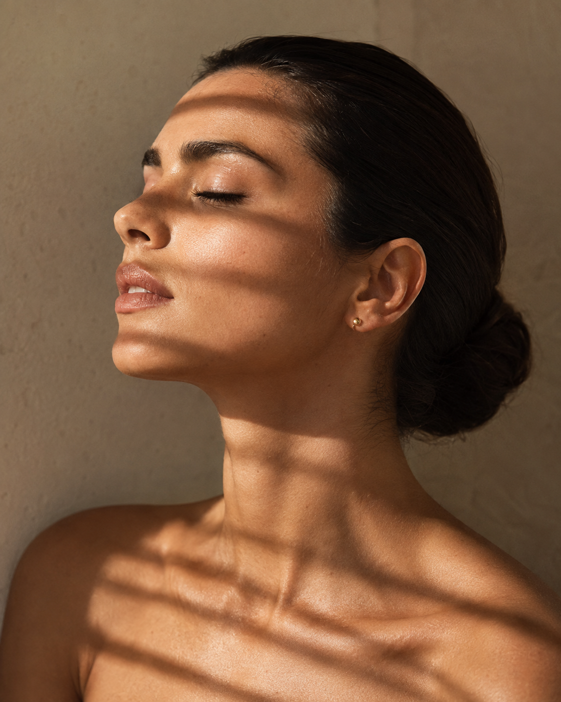 A woman with closed eyes and dark hair styled in a low bun, showing her profile and shoulder against a beige background with shadows from blinds.