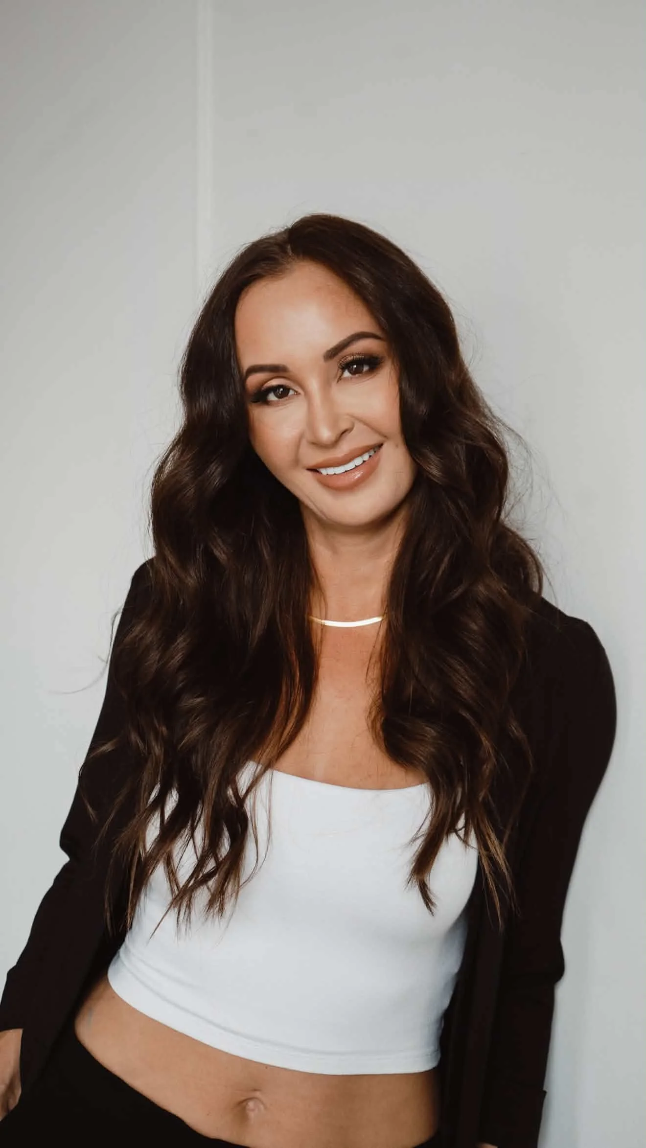 A woman with long, wavy brown hair, makeup, and a smile, wearing a black blazer over a white crop top and a simple necklace, standing against a plain white wall.