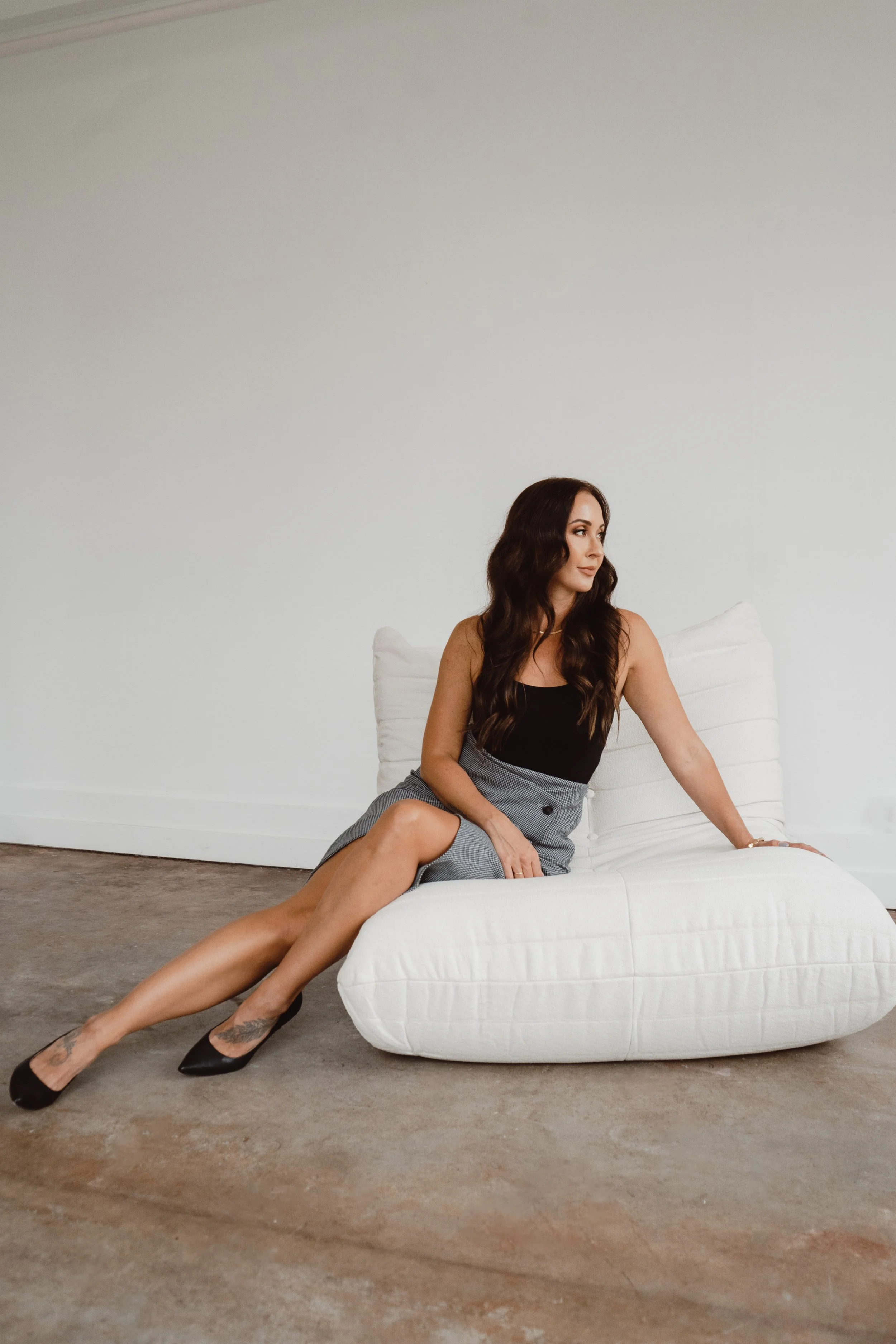 A woman with long dark hair sitting on a white couch in a minimalist room, wearing a black top, gray skirt, black heels, and looking to her left.