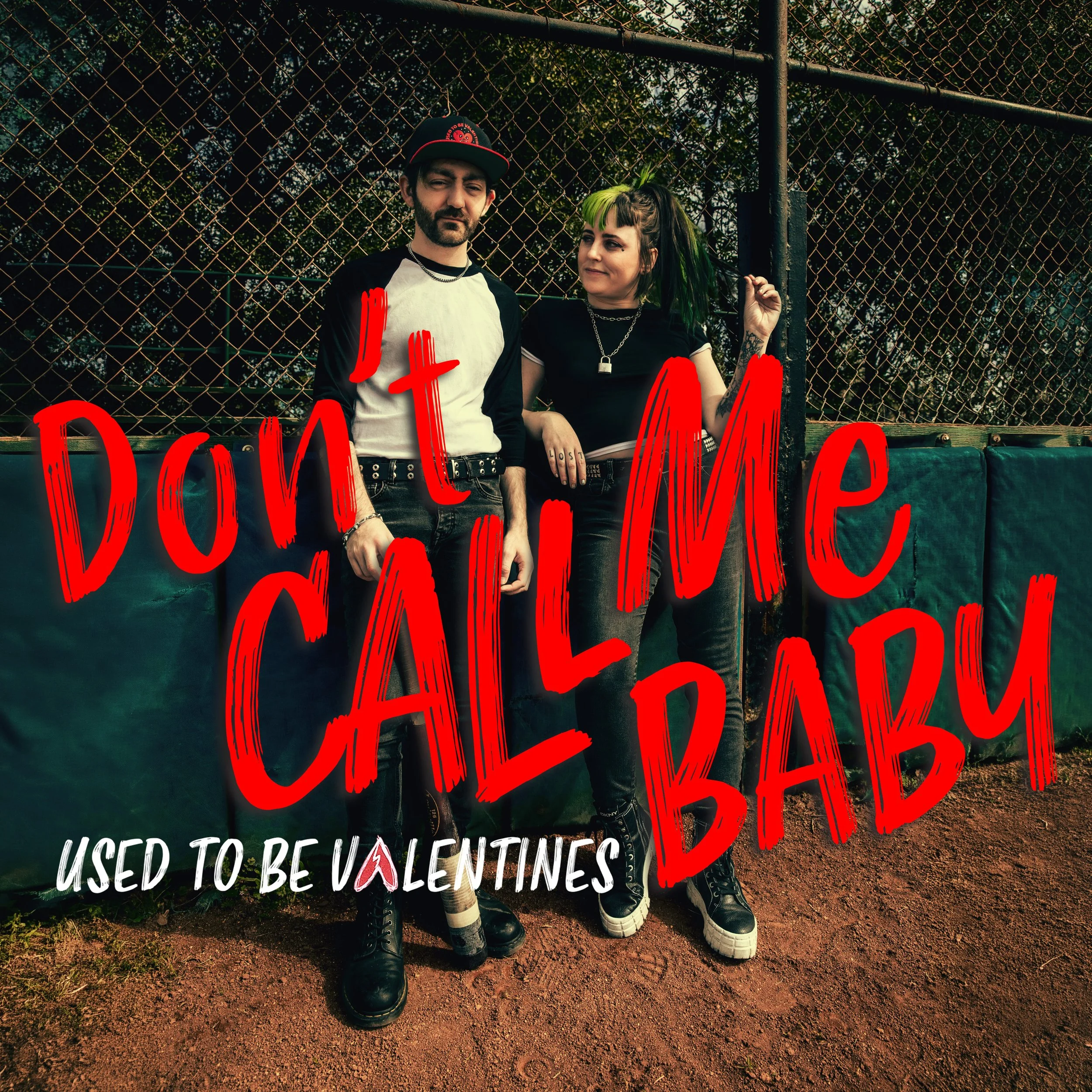 Two young people, one male with a beard and the other female with green and black hair, standing together behind a sign on a chain-link fence. The sign has red and white text that reads 'USED TO BE VALENTINES' and other partially visible words.