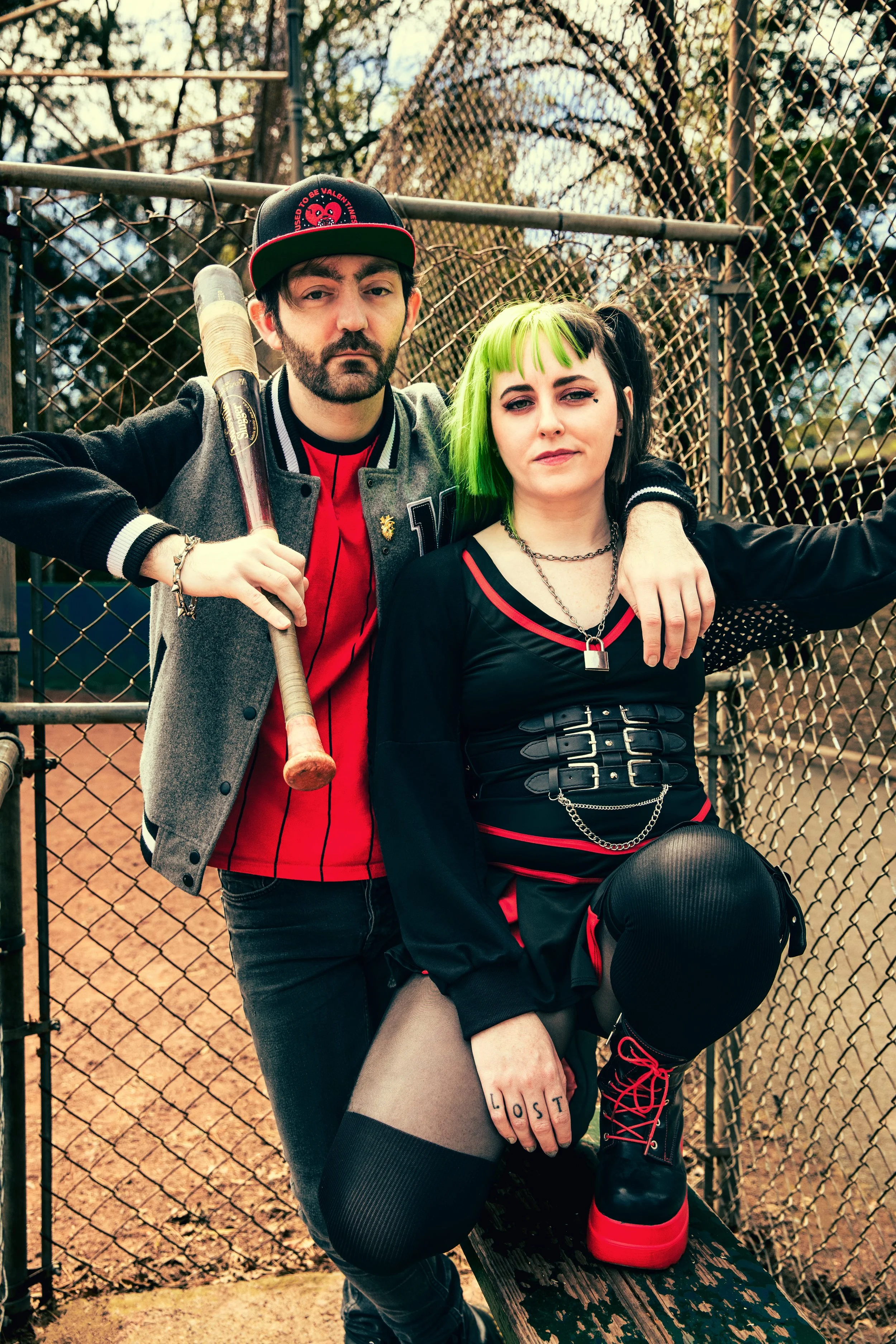 A man with a beard in a red baseball cap and a woman with green hair and a piercing, sitting on a park bench in front of a chain-link fence with trees in the background. The man holds a baseball bat resting on his shoulder, and the woman sits with her arm around him, wearing a black and red dress, black boots with red laces, and fishnet stockings. The woman’s hand on her knee has the word "LOST" tattooed on her fingers.