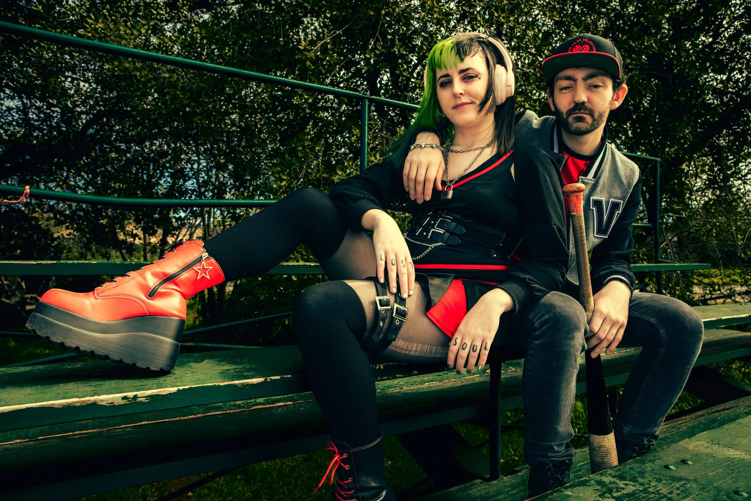 Two young adults sitting on a park bench surrounded by greenery. The woman has green and black hair, wears headphones, black and red clothing, and bright red platform shoes. The man has dark hair, a beard, and wears a baseball cap, a varsity jacket, and dark clothing.