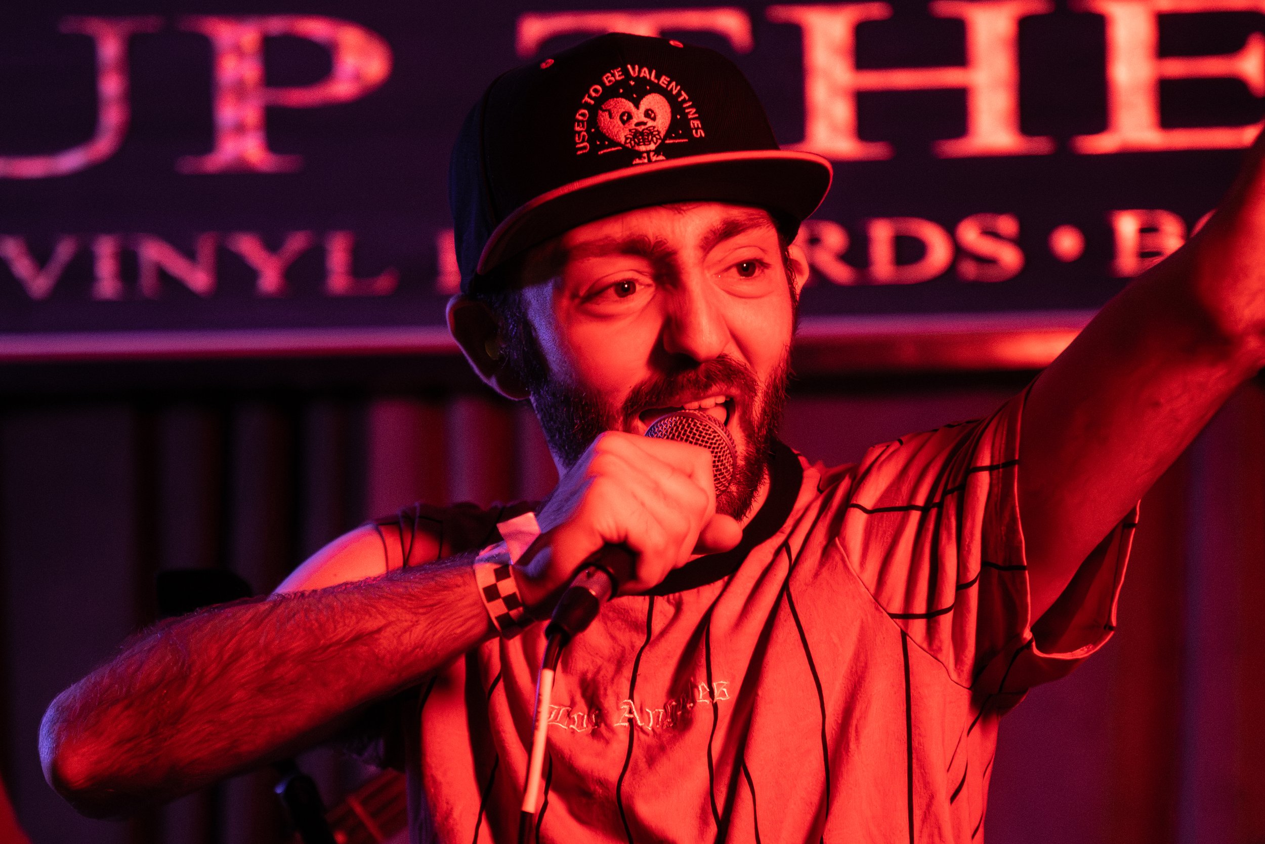 A man singing into a microphone on stage with a dark background illuminated by red lighting, wearing a black cap with a skull and heart design and the words 'Used to Be Valentines.'