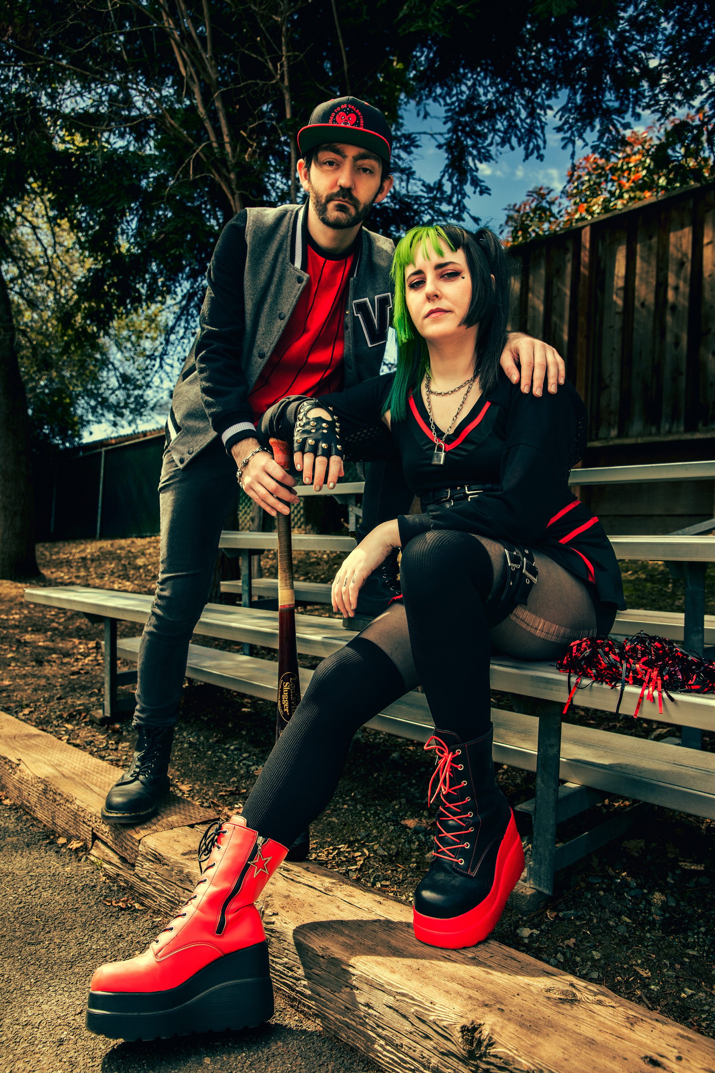 A man and woman dressed in alternative fashion sitting on a park bench outdoors during daytime. The woman has green and black hair, multiple facial piercings, and wears black and red platform boots, a black dress with red accents, and fishnet stockings. The man has a beard, wears a gray letterman jacket, red shirt, and a black cap. They pose with serious expressions, with the woman seated and the man leaning on her shoulder, holding a baseball bat.