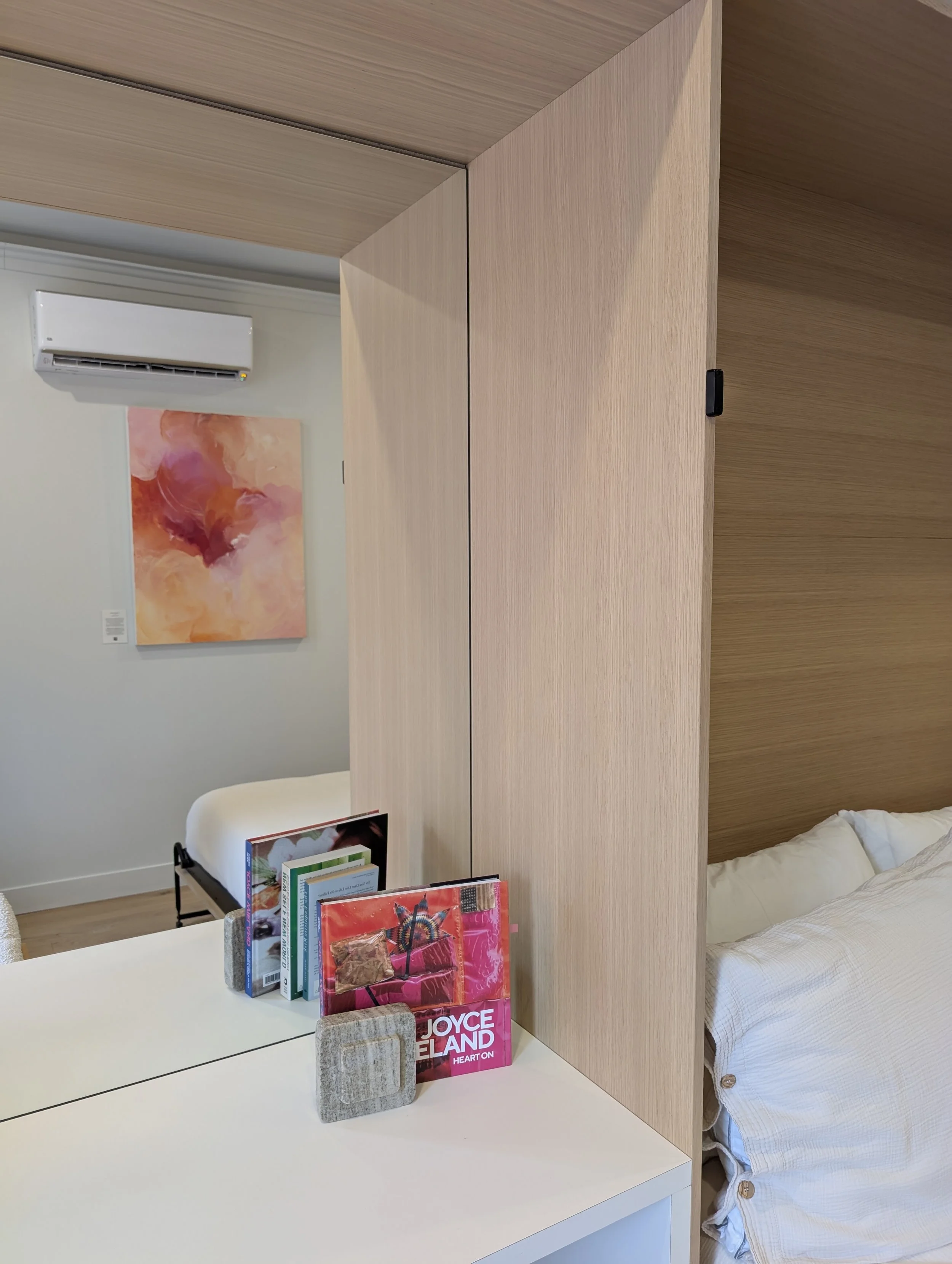 A bedroom with a wooden divider, a bed with white linens, a white table with books and a Joyceeland Heart on tote bag, a painting with pink and orange hues, and an air conditioning unit on the wall.