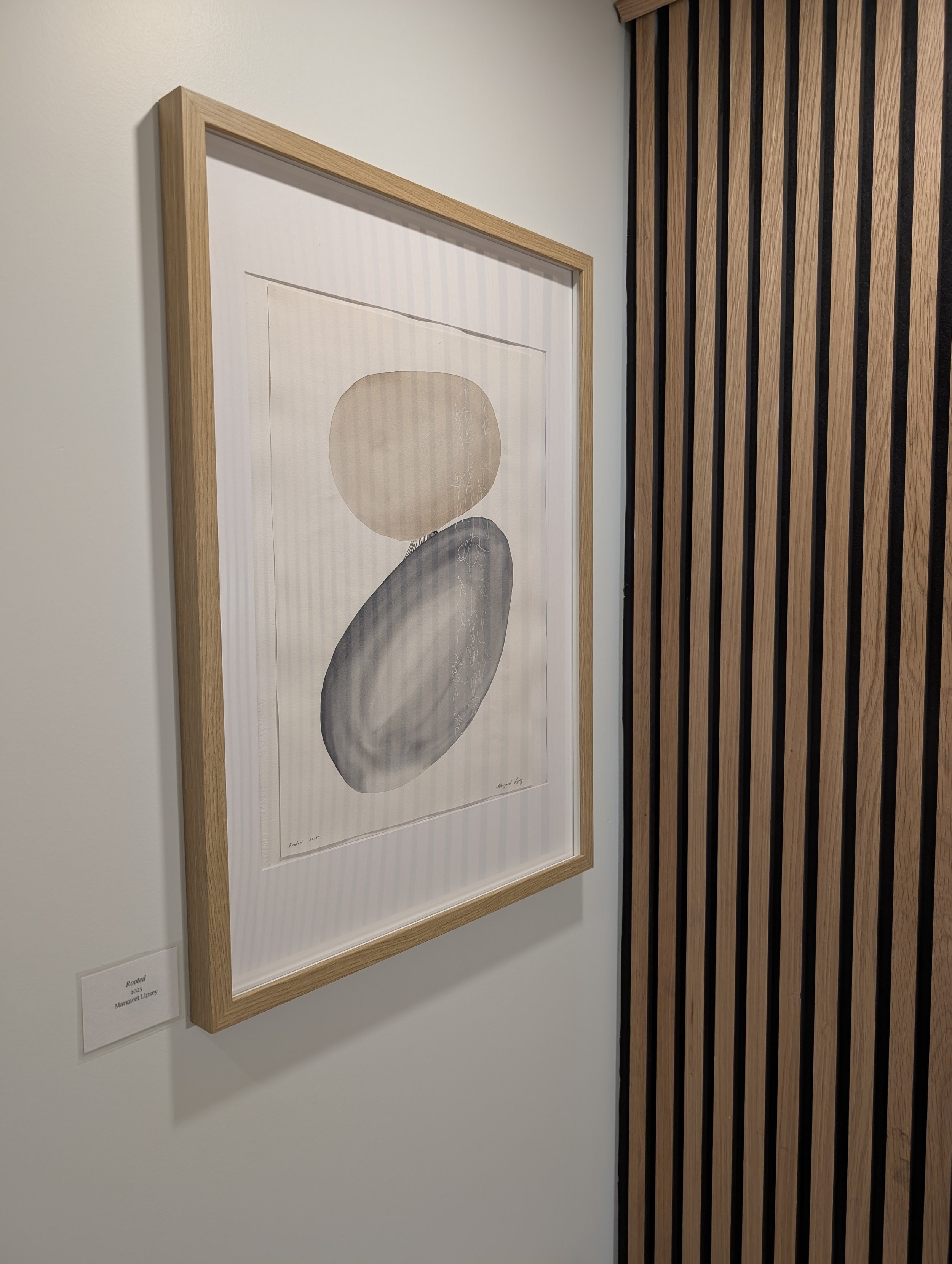 Abstract artwork of two overlapping circles, one beige and one gray, with a shadow pattern, framed and displayed on a white wall with a wooden panel on the side.