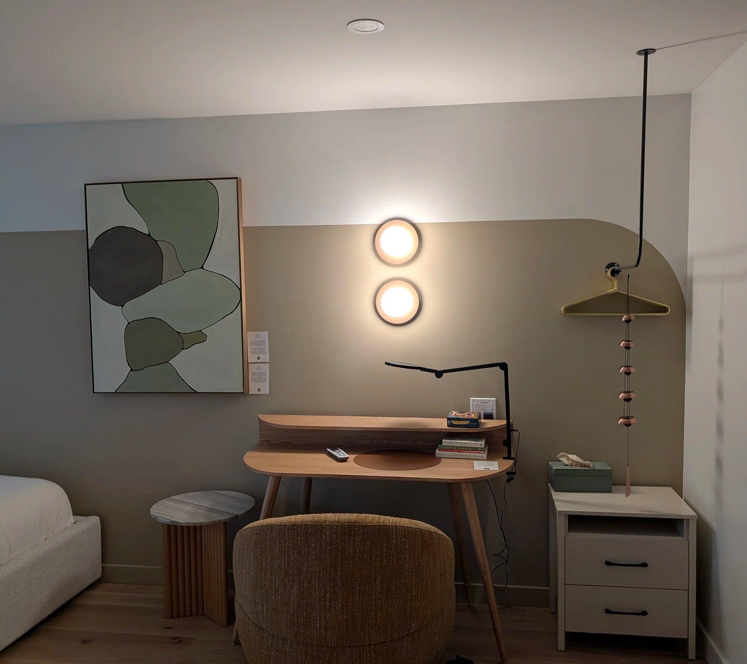 A cozy interior with a wooden desk, beige chair, small side table, abstract wall art, two round wall lights, modern hanging lamp, and a small cabinet.