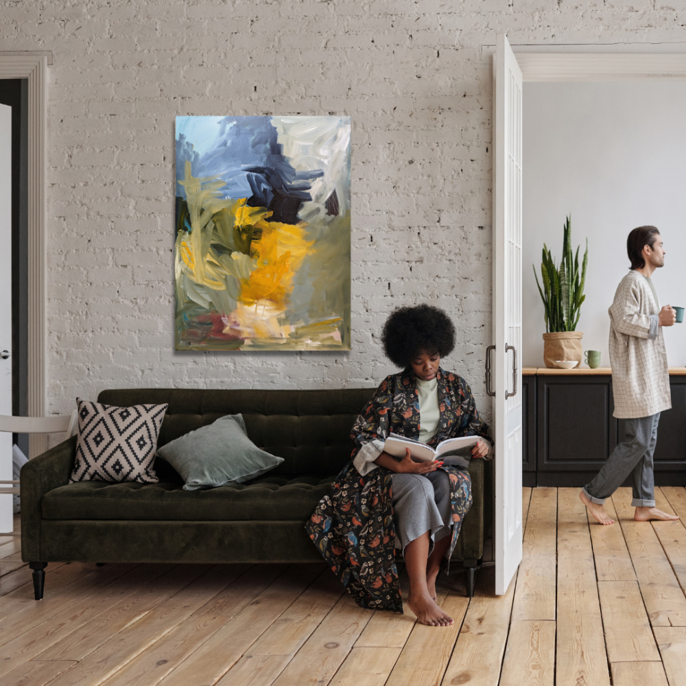A woman with afro hair sitting on a dark green sofa reading a book, next to two pillows, one with a geometric pattern and one plain. Behind her is a white brick wall with an abstract painting. To the right, a woman with short hair in pajamas is walking and holding a mug in a kitchen area with black cabinets, a potted plant, and a wooden floor.