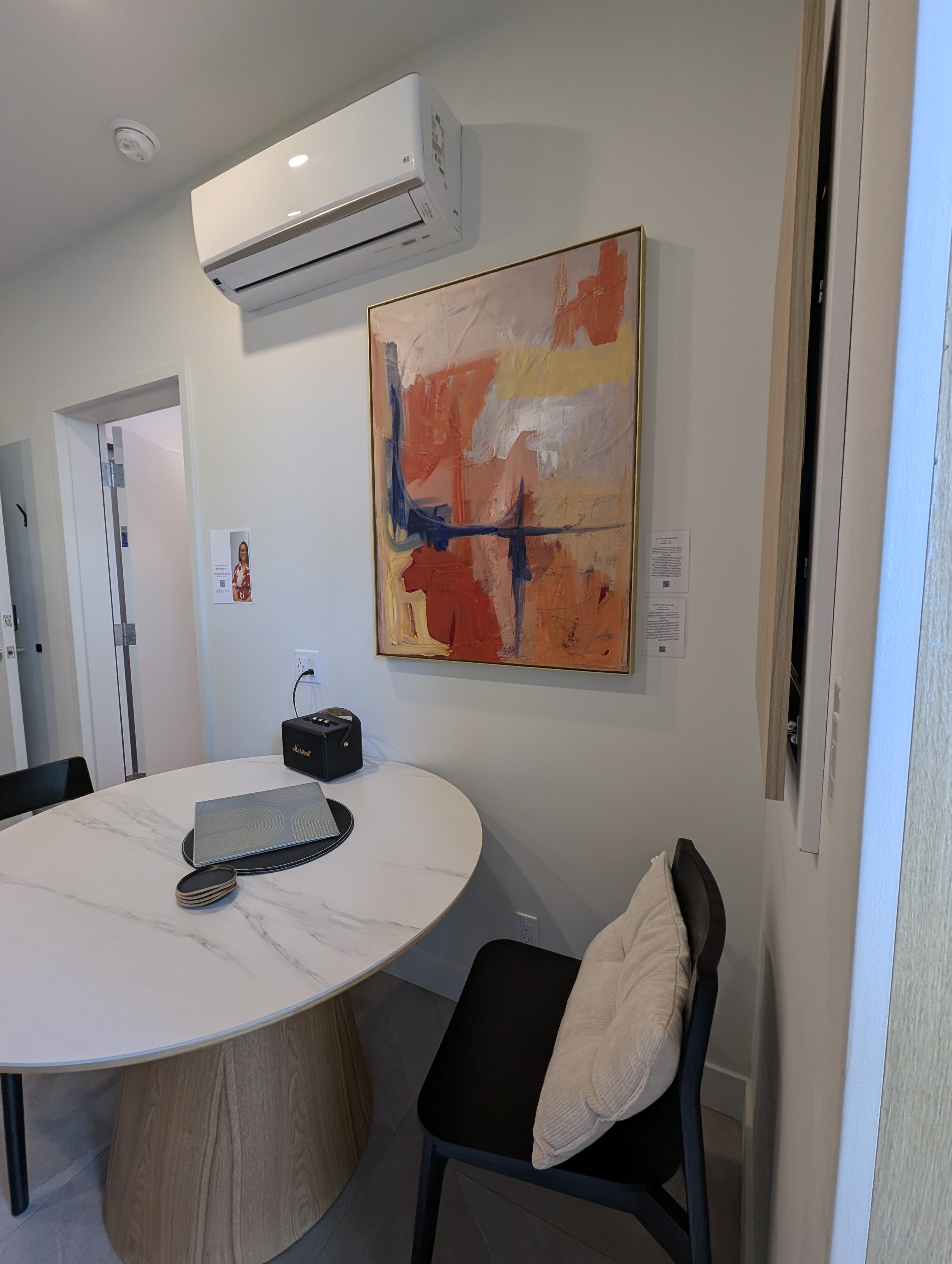 Modern dining room with a white marble oval table, a black chair with a beige cushion, colorful abstract wall art, an air conditioner, and a Marshall speaker.