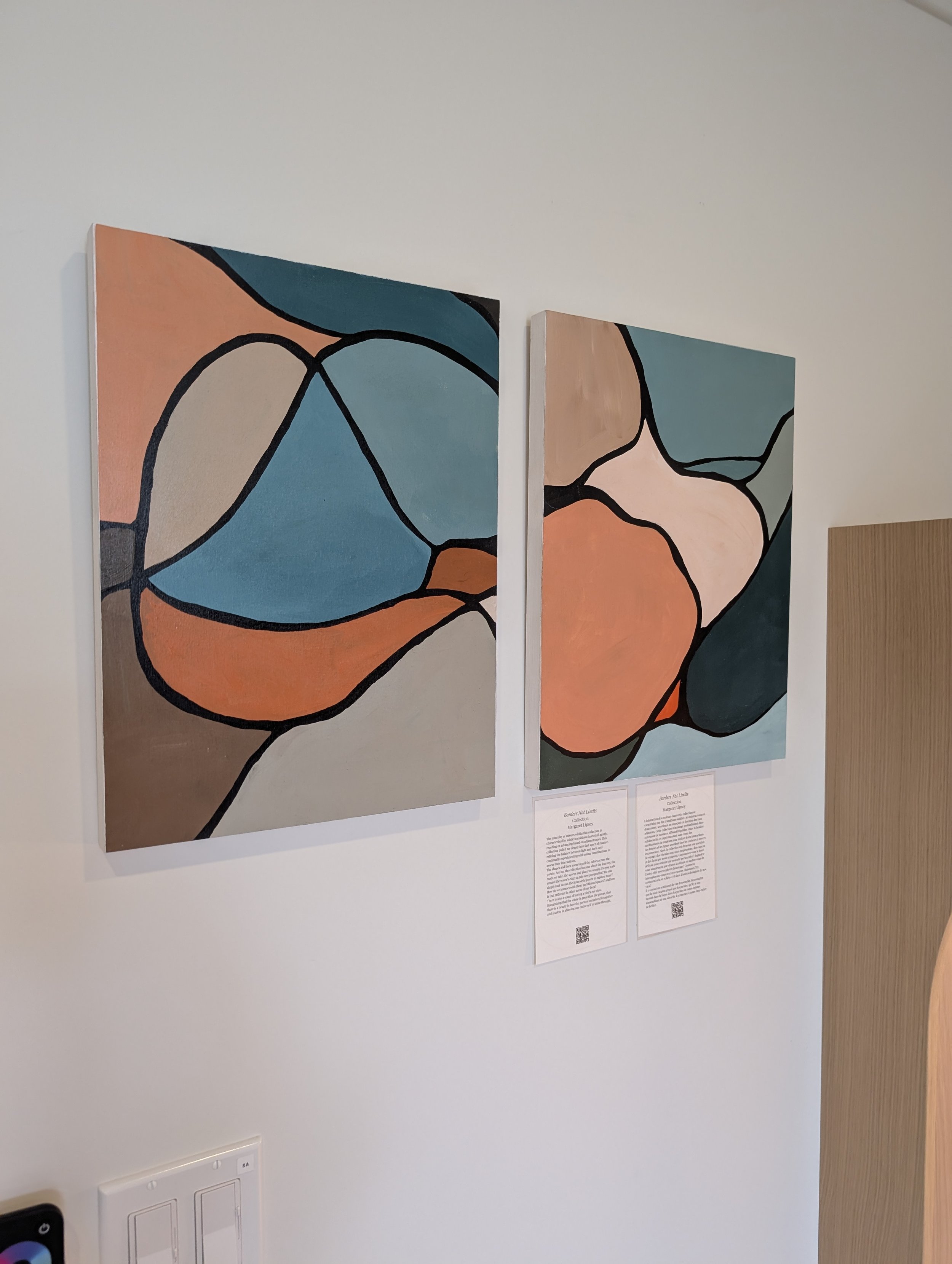 Two abstract landscape paintings with bold black outlines and soft colors, hanging on a white wall, with informational placards below them.