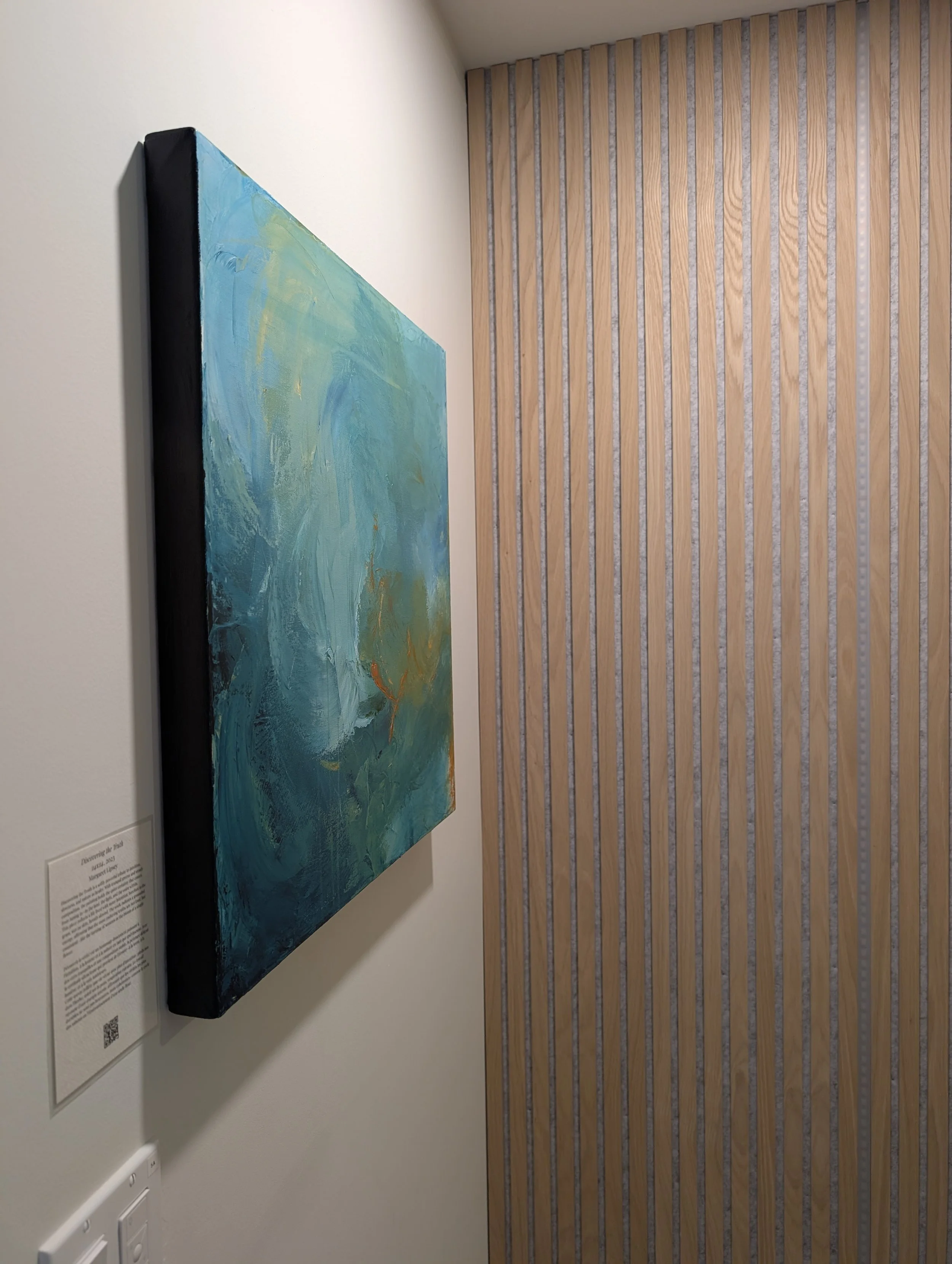 An abstract painting with swirling blue, green, and yellow colors hanging on a white wall in an art gallery.