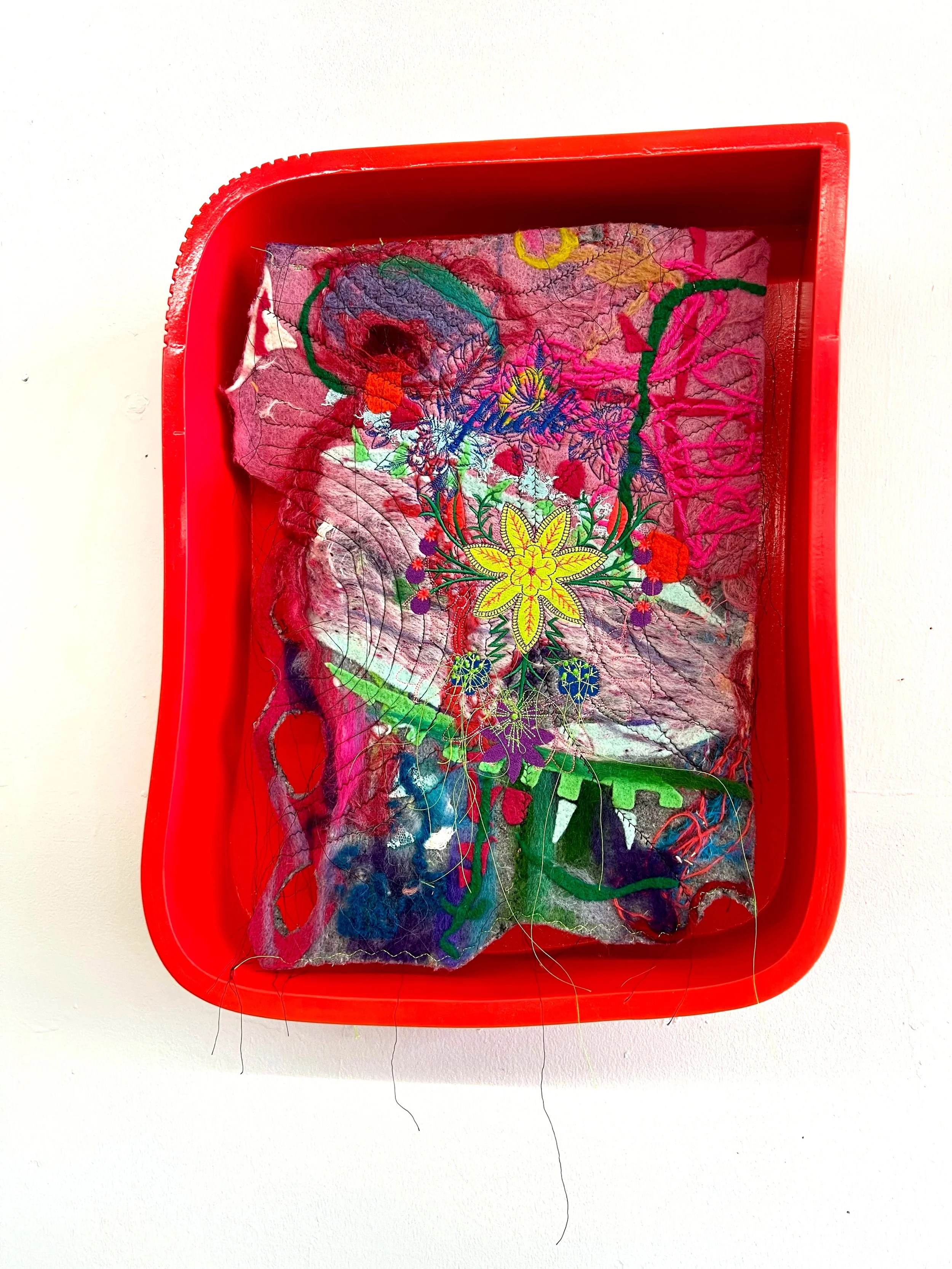Red plastic tray holding a piece of fabric with colorful embroidery and torn sections.