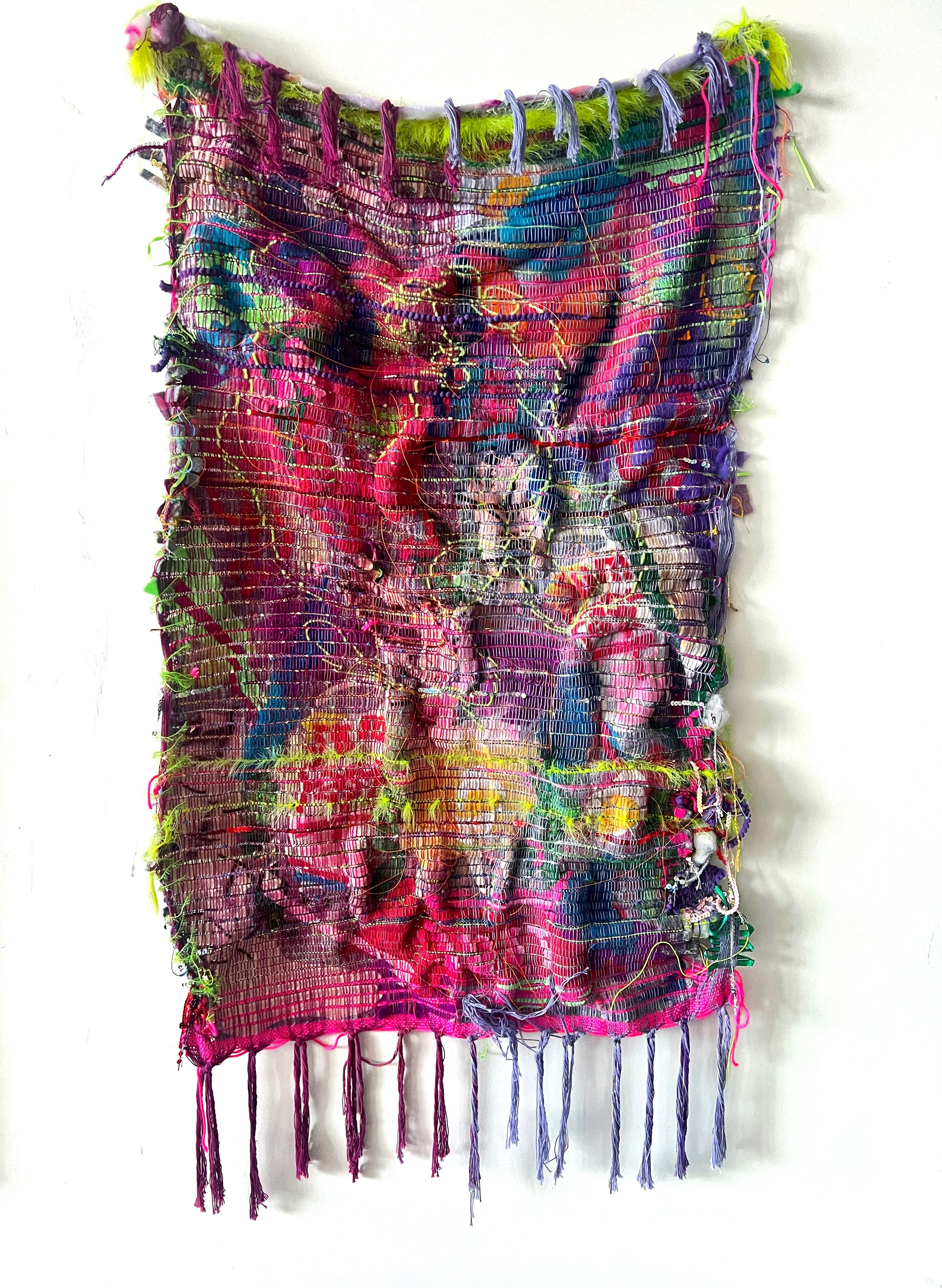 A colorful, textured textile art piece with various fabric, threads, and embellishments sewn onto a mesh or net background, hanging vertically on a white wall.