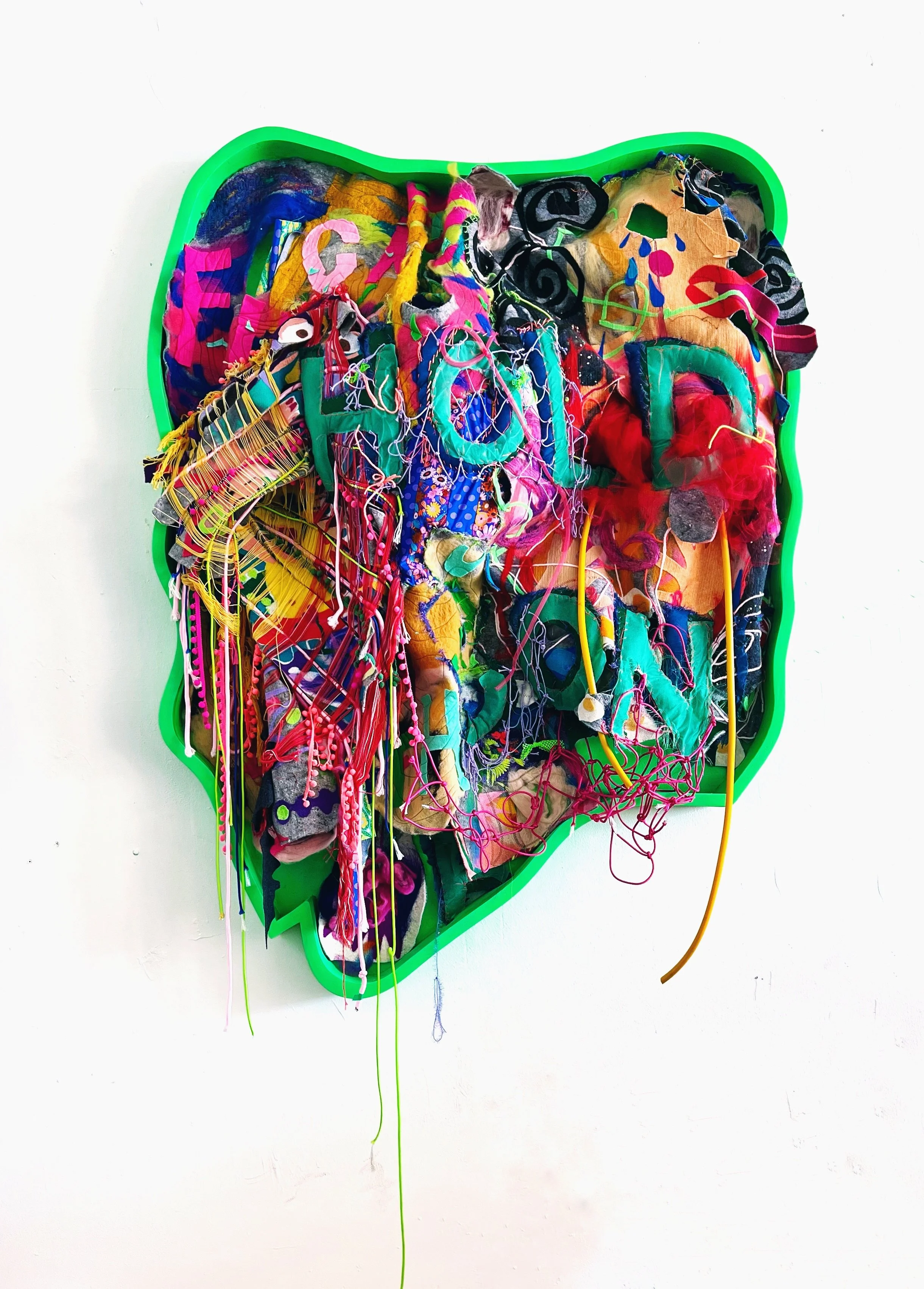 Mixed media artwork composed of colorful fabric, yarn, and thread inside a neon green heart-shaped frame, attached to a white wall.