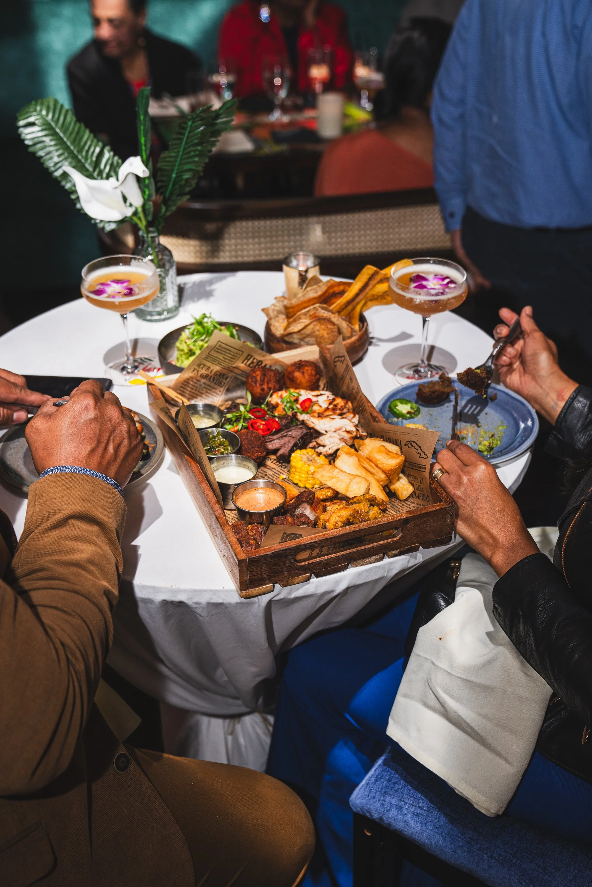 People dining at a restaurant, sharing a large tray of assorted foods including meats, fries, and vegetables, with two cocktails on the table and a decorative plant in the background.