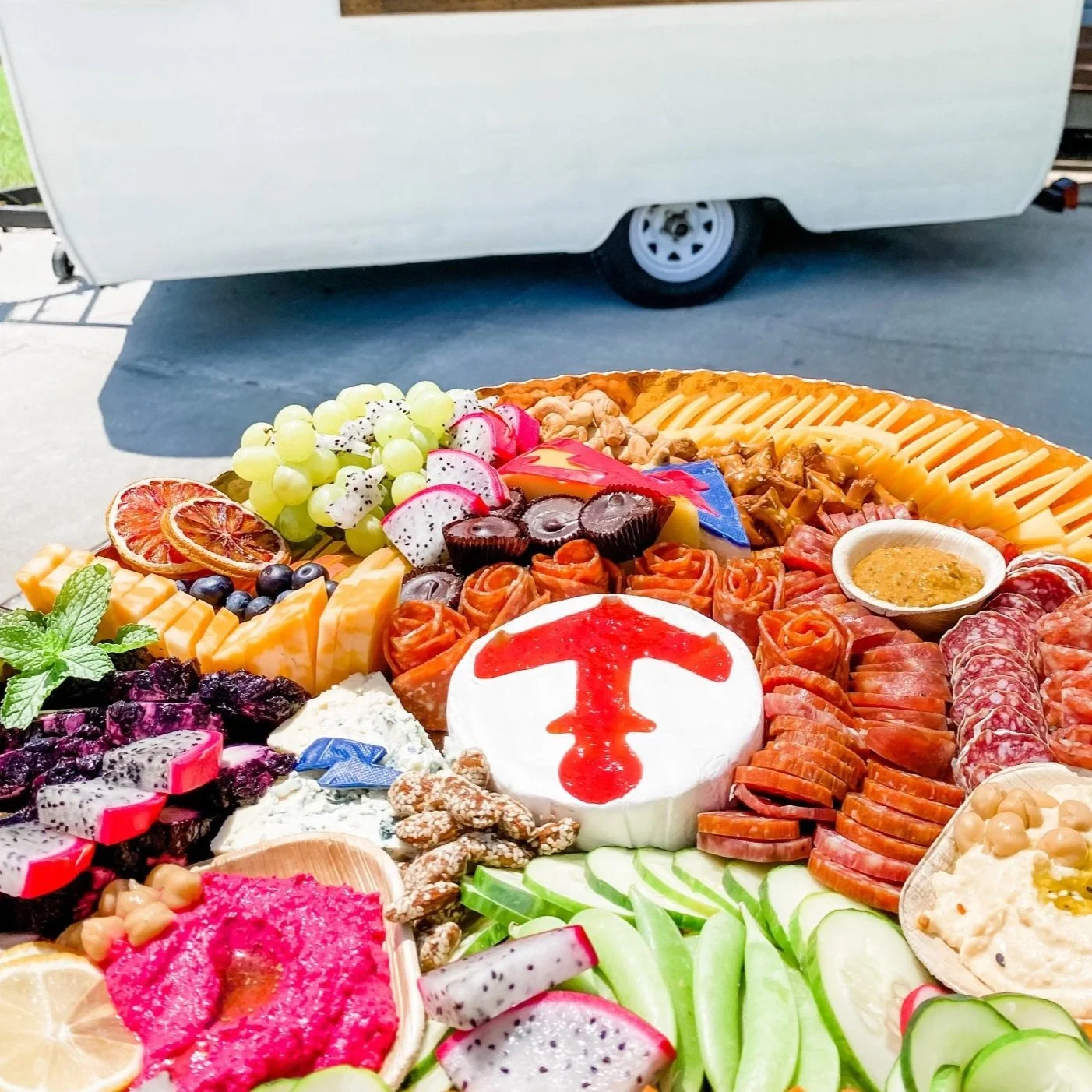 Colorful charcuterie board with various meats, cheeses, fruits, crackers, vegetables, nuts, and dips arranged on a circular plate. There is a round cheese with a red symbol on top at the center.
