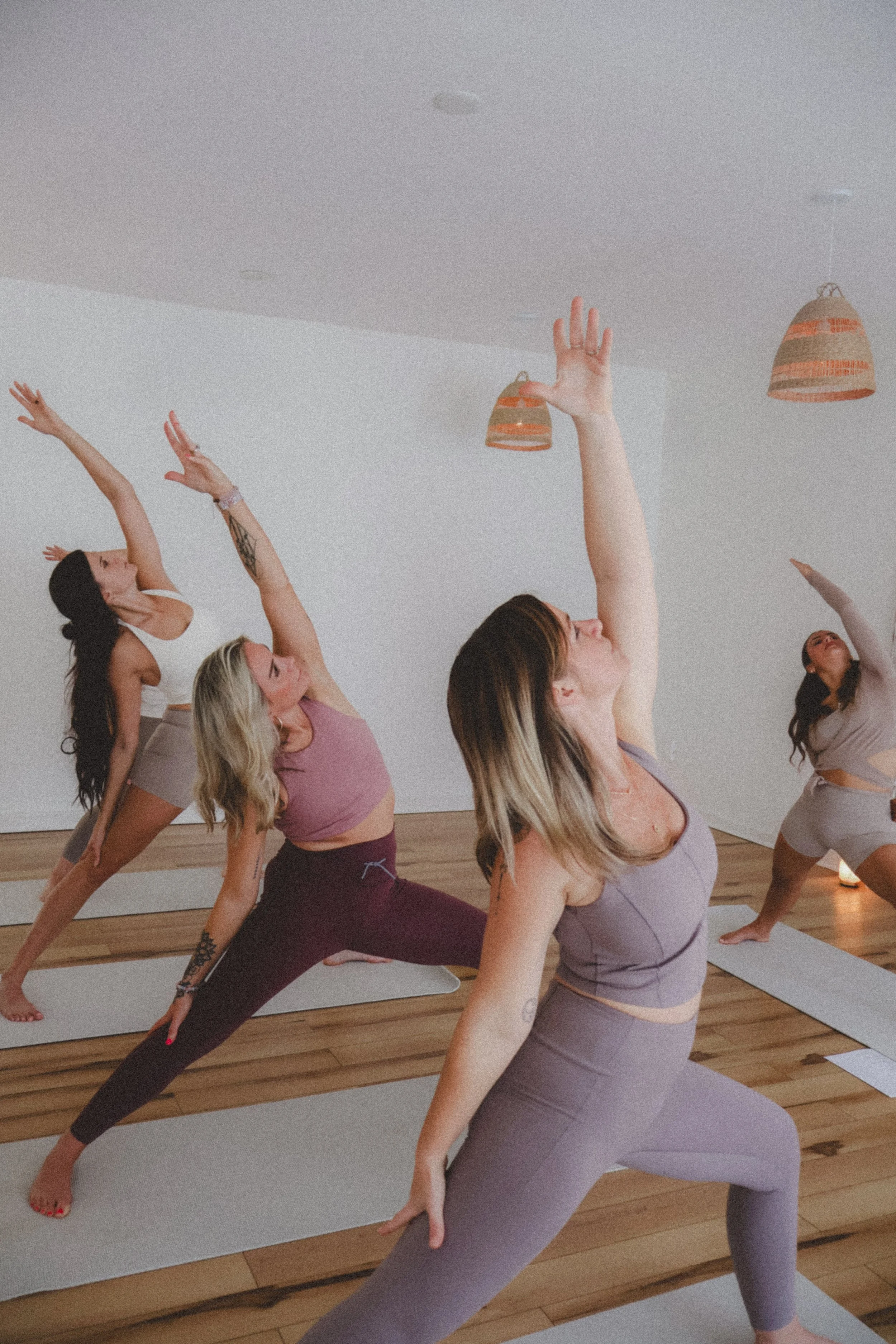 Group of women practicing yoga in a studio, with wooden floors and natural light, performing a warrior pose on yoga mats.