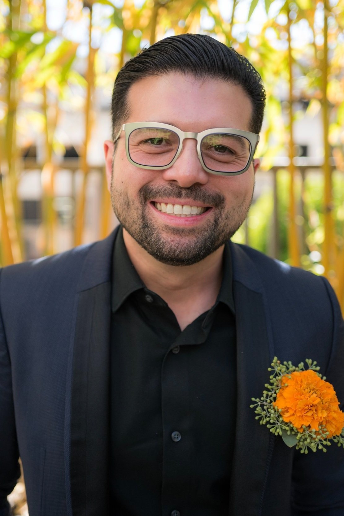 A smiling man with glasses, wearing a dark suit and black shirt, with a bright orange flower on his left lapel, outdoors with yellow and green foliage in the background.