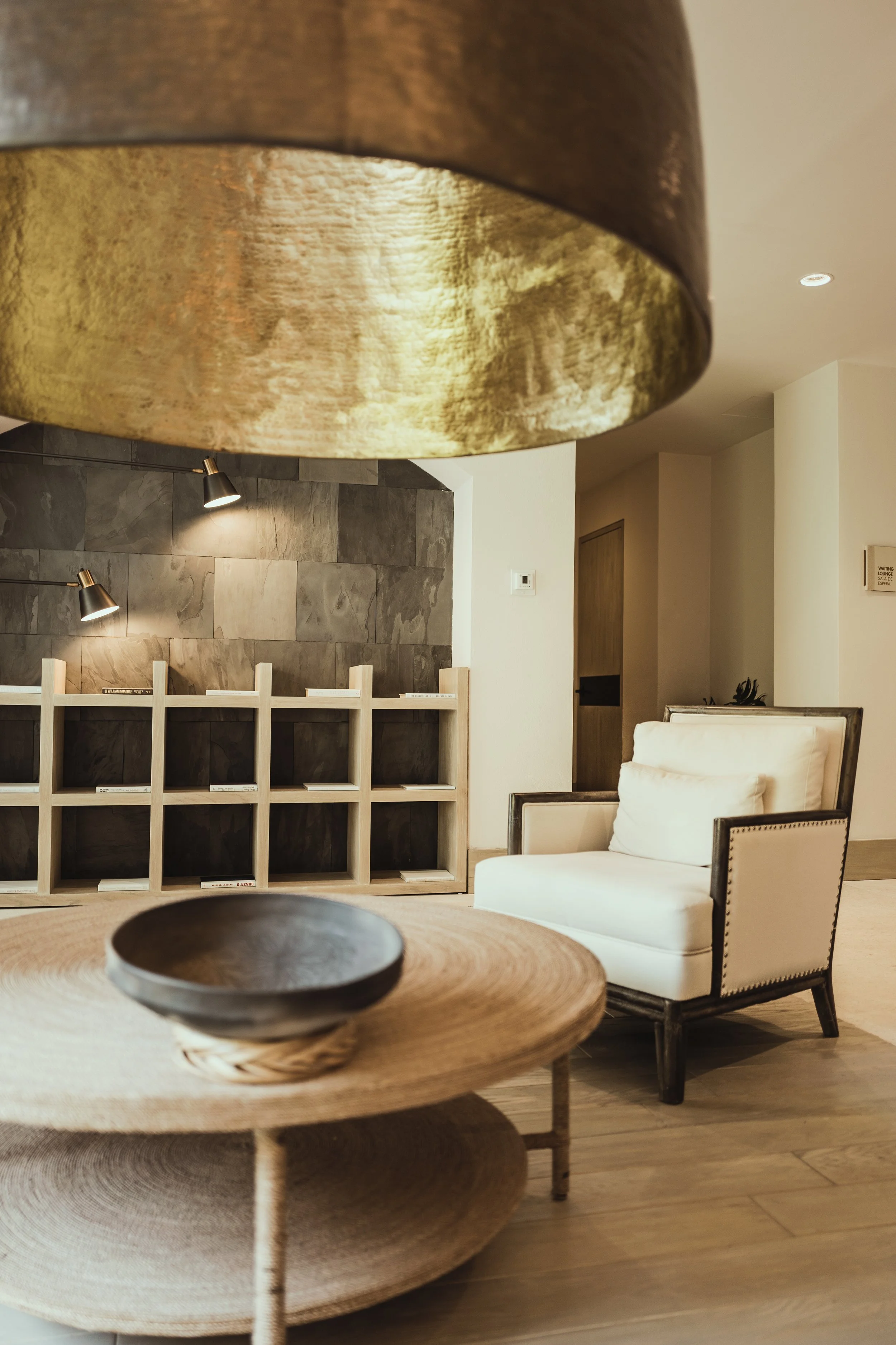 Interior of a modern room featuring a large, round, metallic gold-colored hanging lamp, a wooden coffee table with a bowl, a white armchair with a pillow, a stone accent wall with two adjustable reading lamps, and a wooden shelf with compartments.