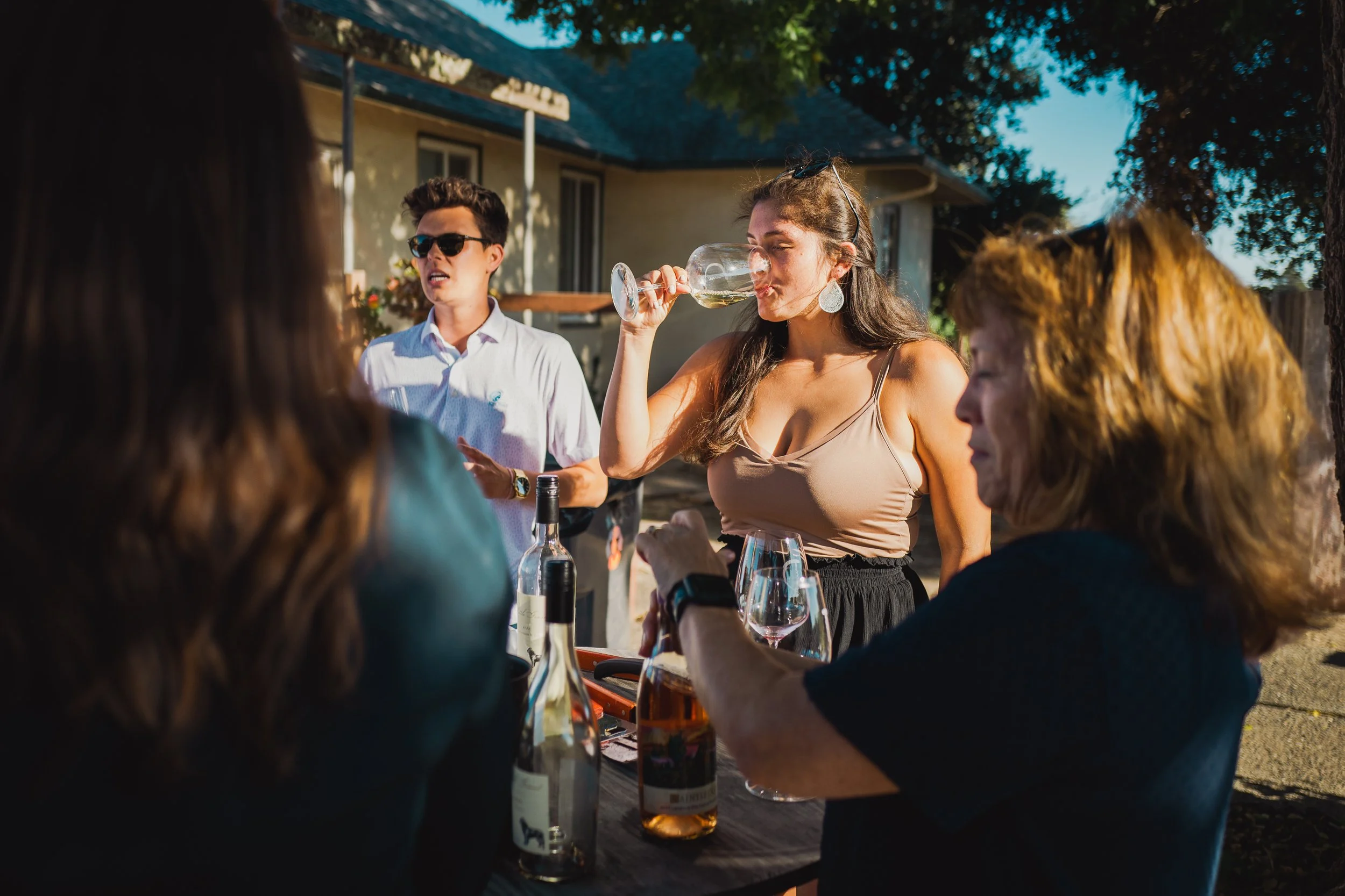 Group of adults sitting outdoors at a sunset gathering, with a woman drinking wine and a young man standing nearby.