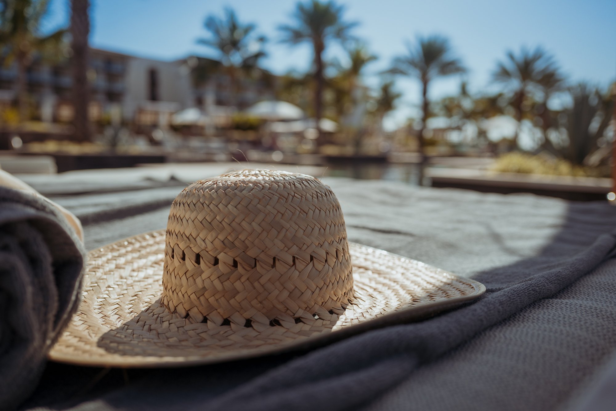 A straw sun hat resting on a beach towel with a resort and palm trees in the background under a clear blue sky.