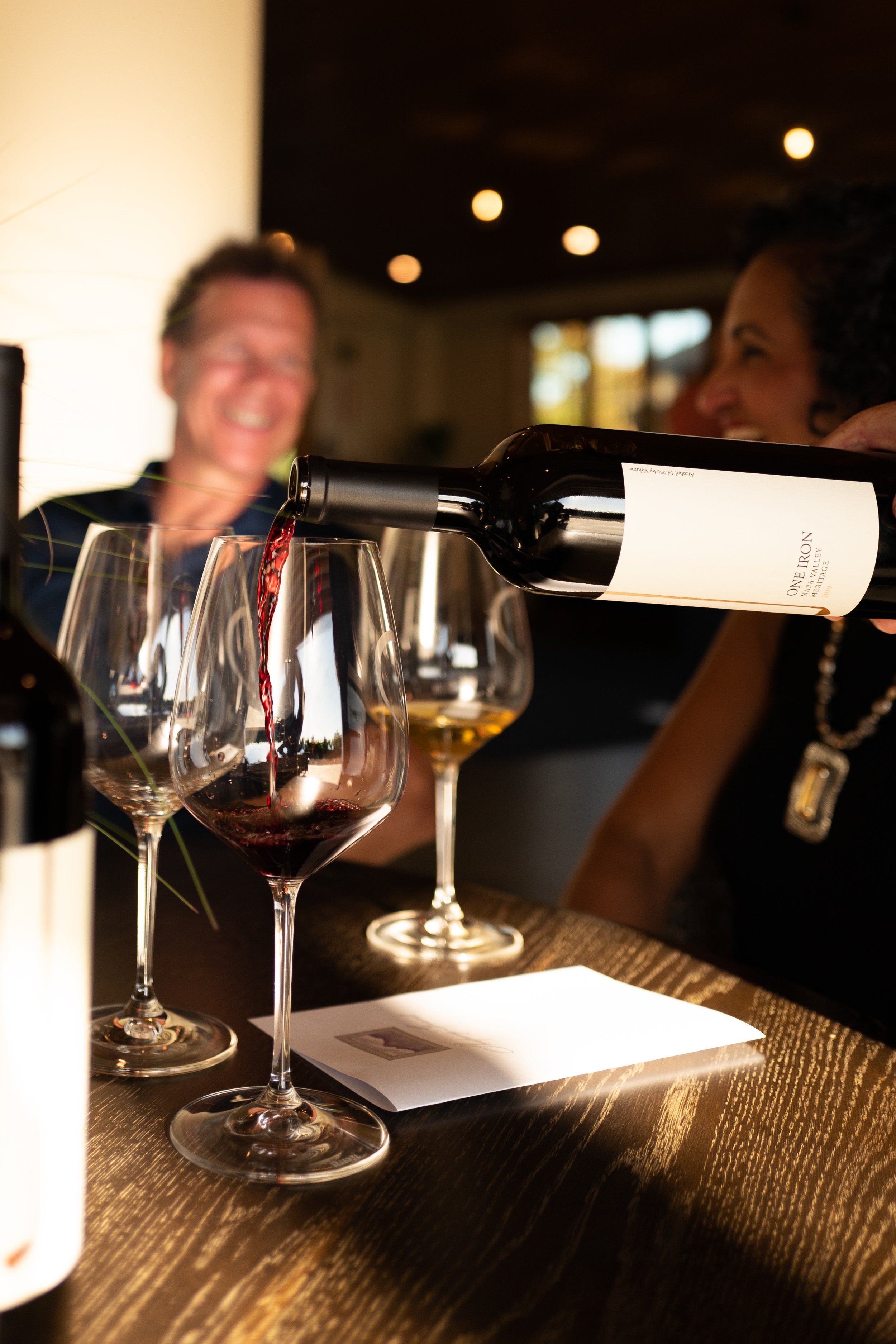 A person is pouring red wine into a glass at a bar, with two other wine glasses and a blurred smiling couple in the background.